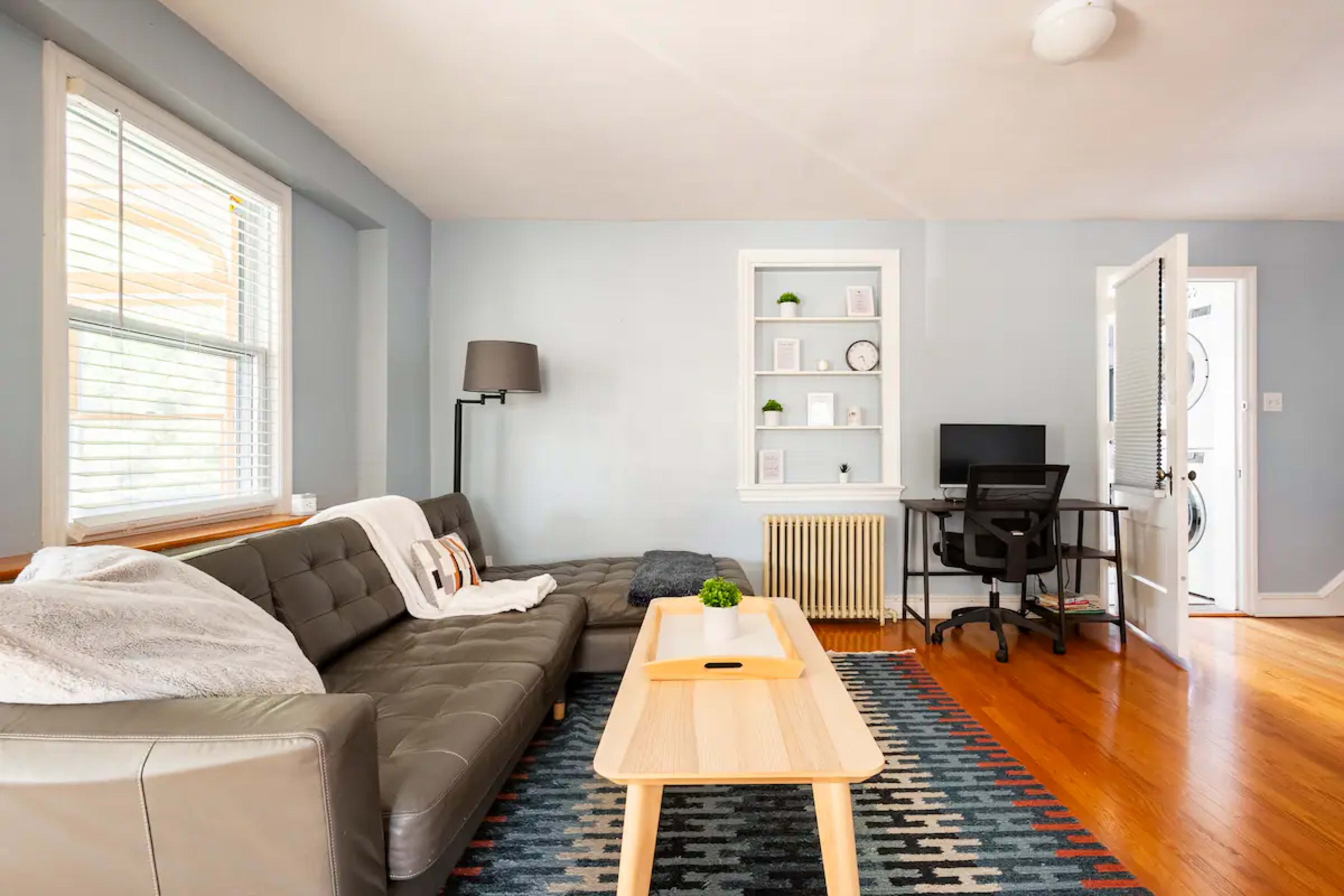 A living room features a gray couch, a wooden coffee table, a chair at a desk, and a shelf with decorative items against light blue walls.