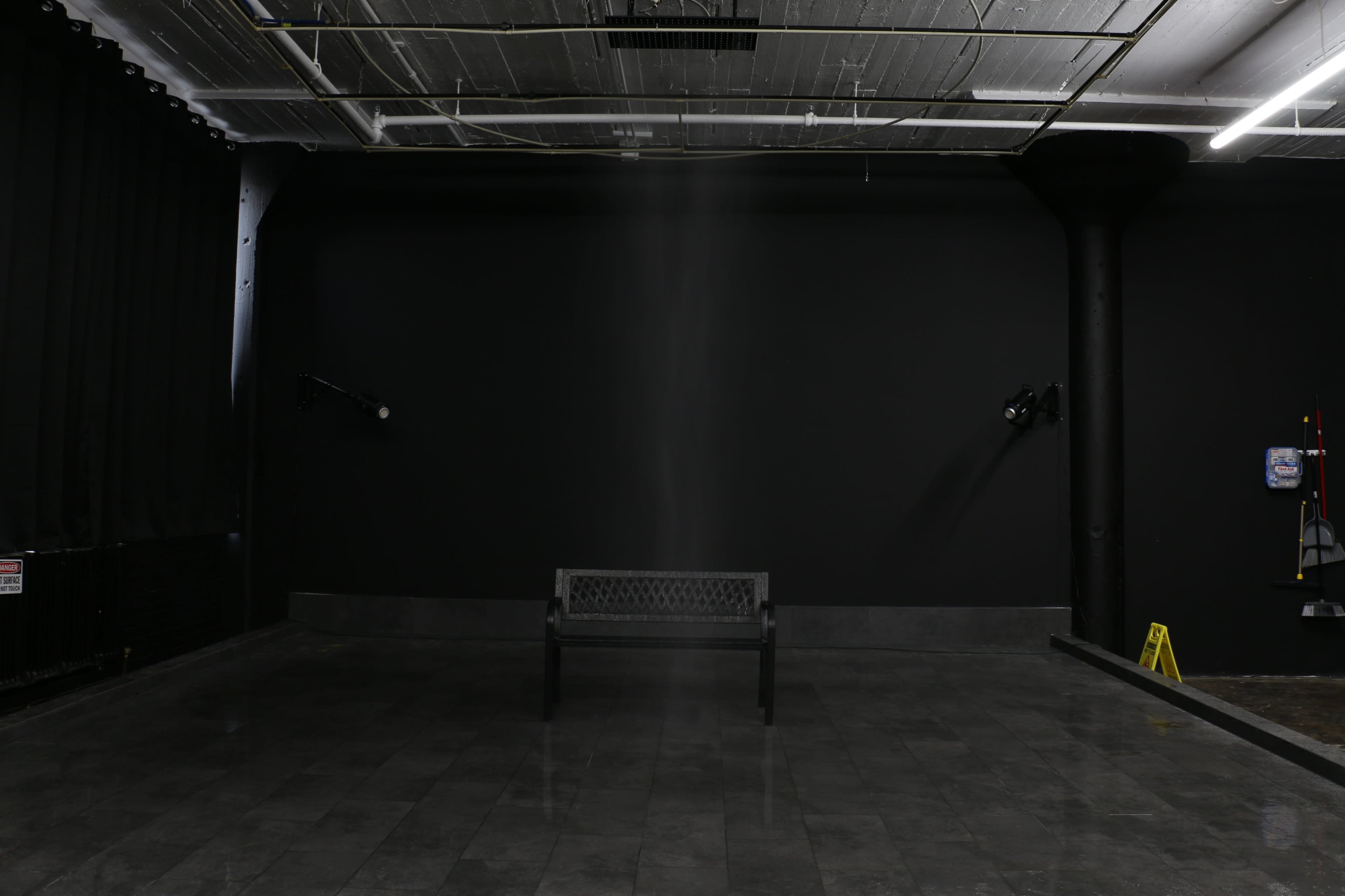 The image shows a sparsely furnished room with a black wall, a single bench in the center, and two overhead lights.