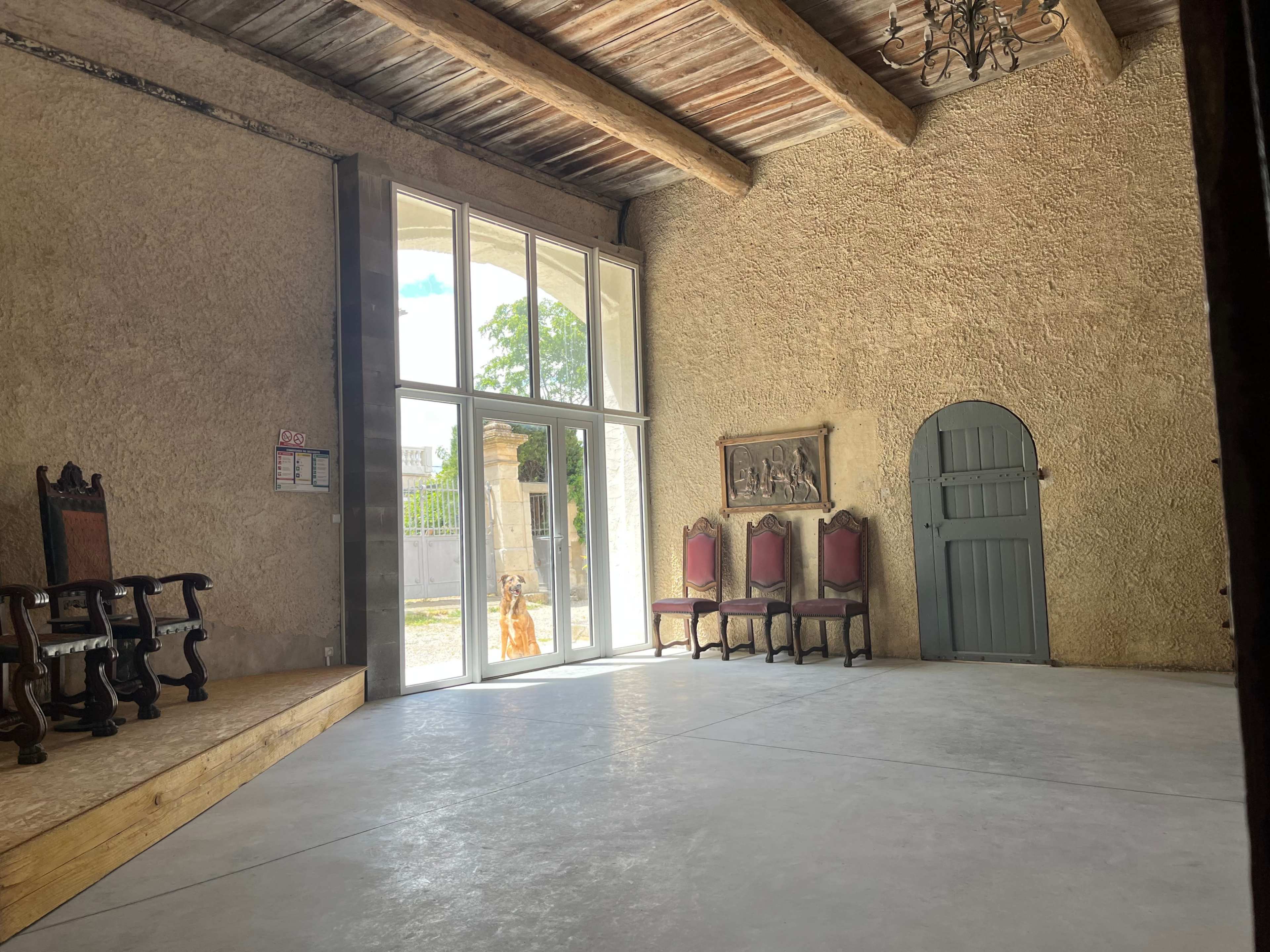 The image depicts a spacious room with a stone-textured wall, a wooden ceiling, and a row of chairs along one side, alongside a large window allowing natural light to enter.