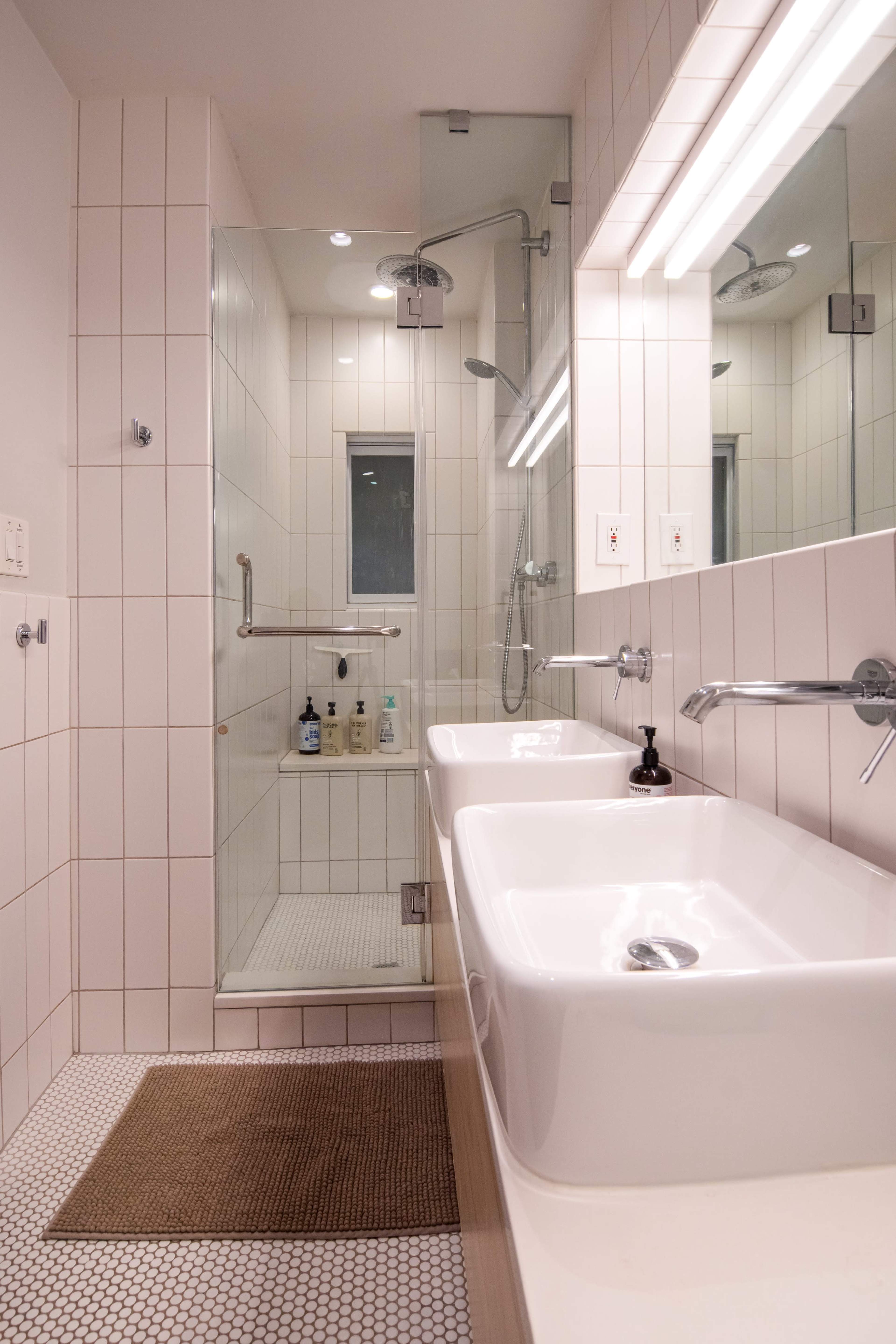 The image shows a modern bathroom featuring dual sinks, a glass-enclosed shower, and a tiled floor with a small rug.