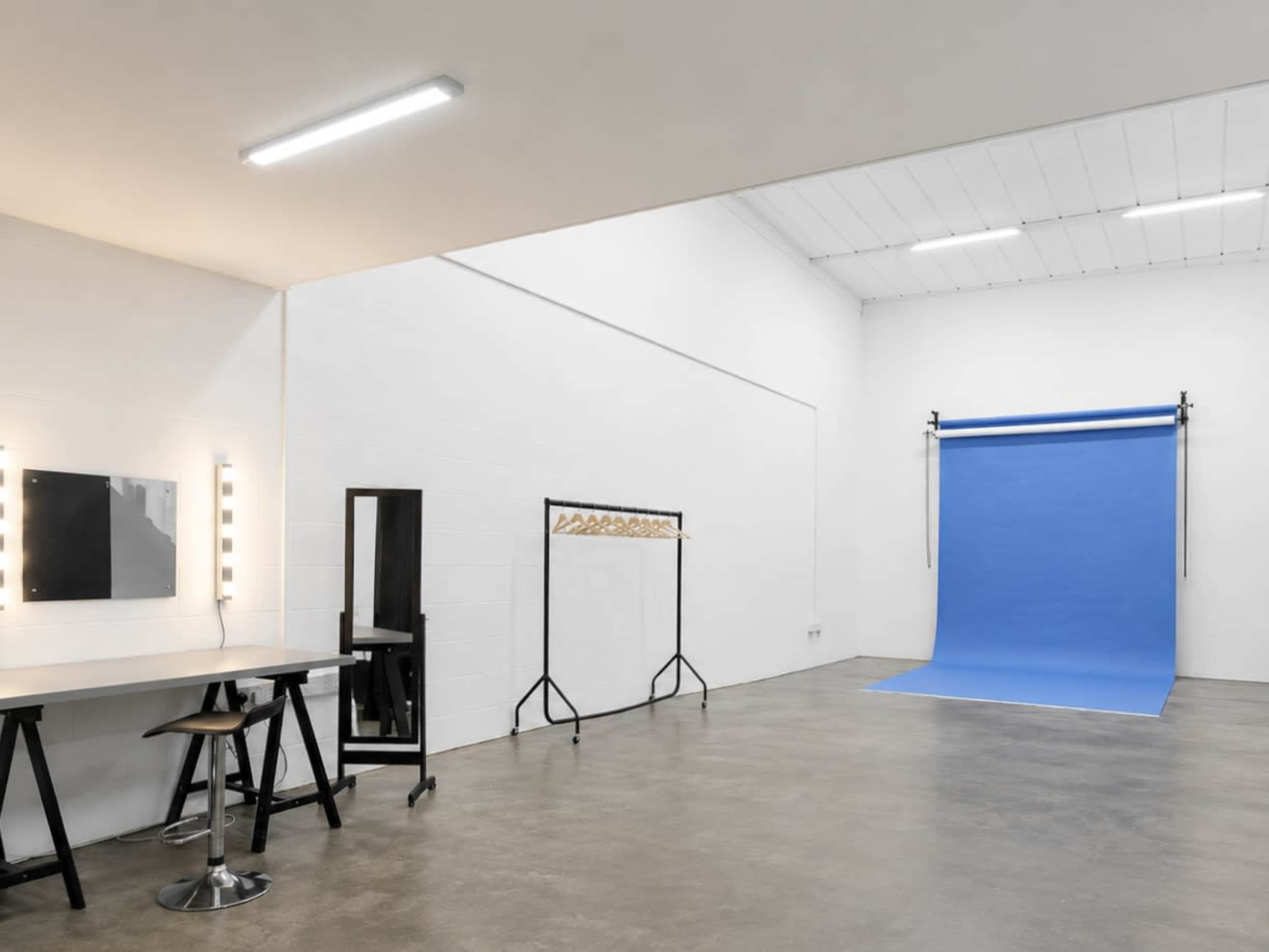 The image shows a well-lit photography studio featuring a blue backdrop, a makeup vanity with lights, and a clothing rack.