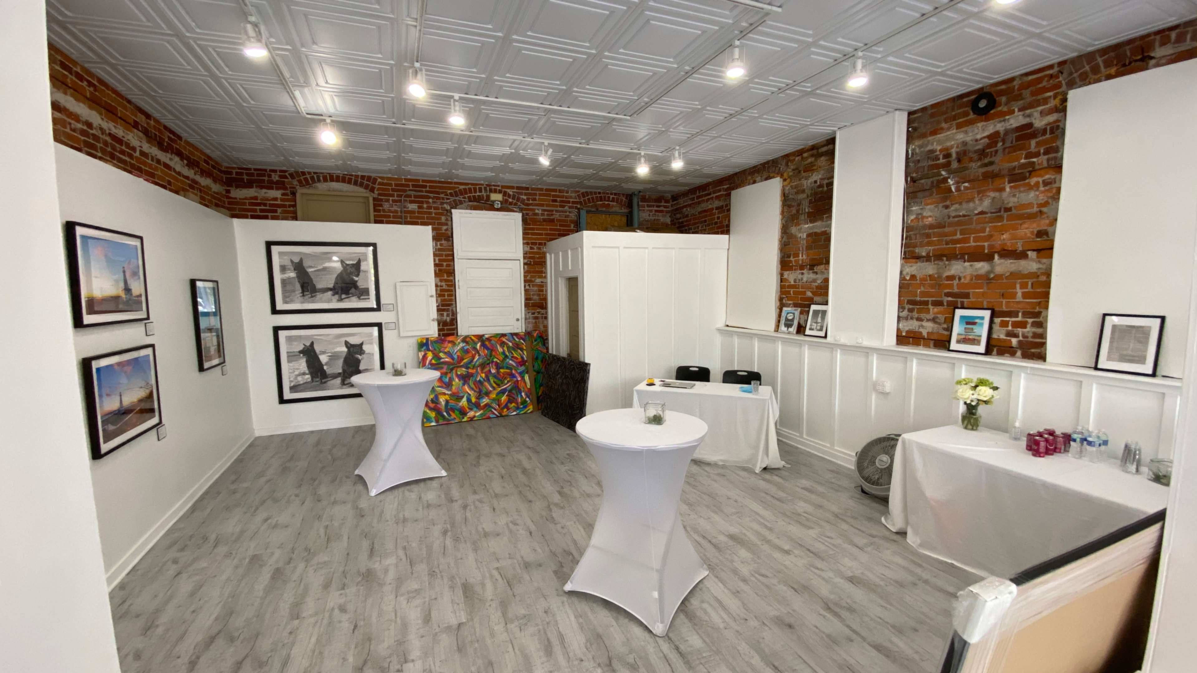 The image shows a spacious room with exposed brick walls, featuring artwork on display and two white cocktail tables set up for an event.