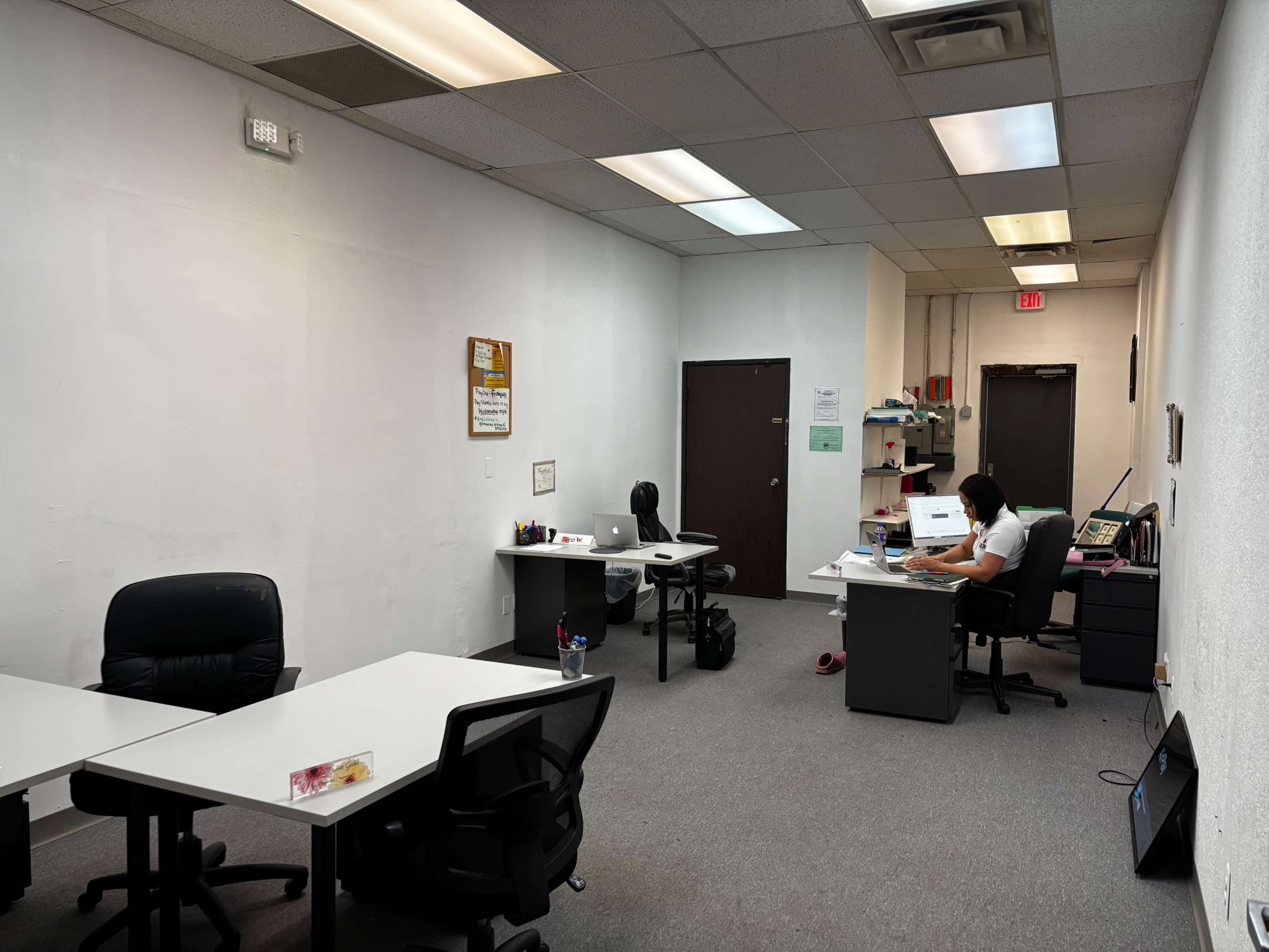 The image shows an office space with two desks and chairs, one occupied by a person working, and a blank wall on the left.
