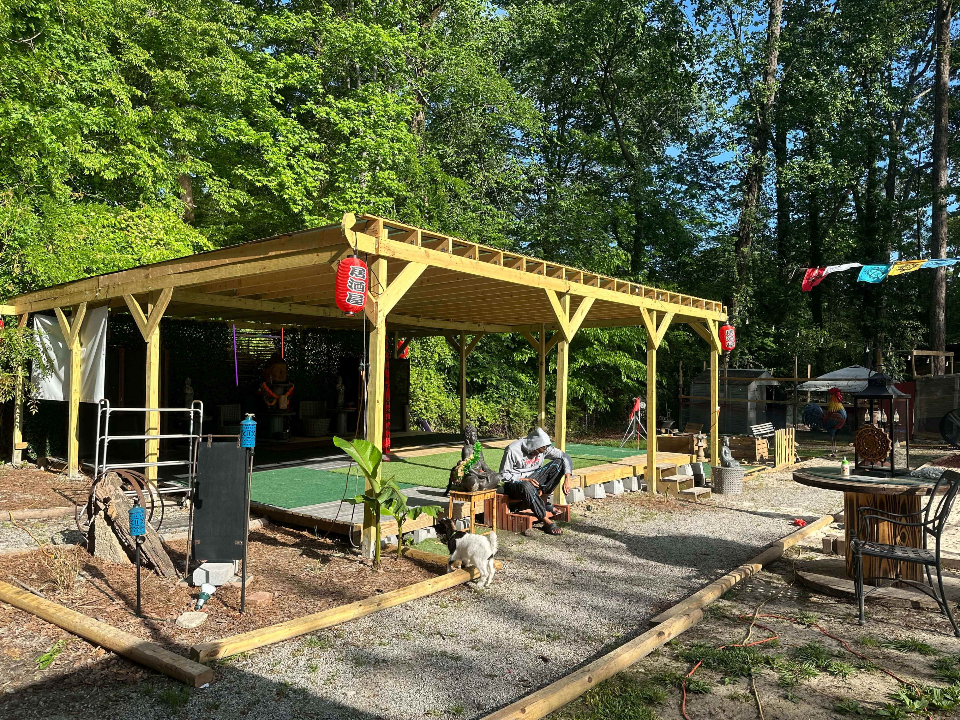 A wooden gazebo structure with benches and a stone pathway is surrounded by green trees and garden elements.