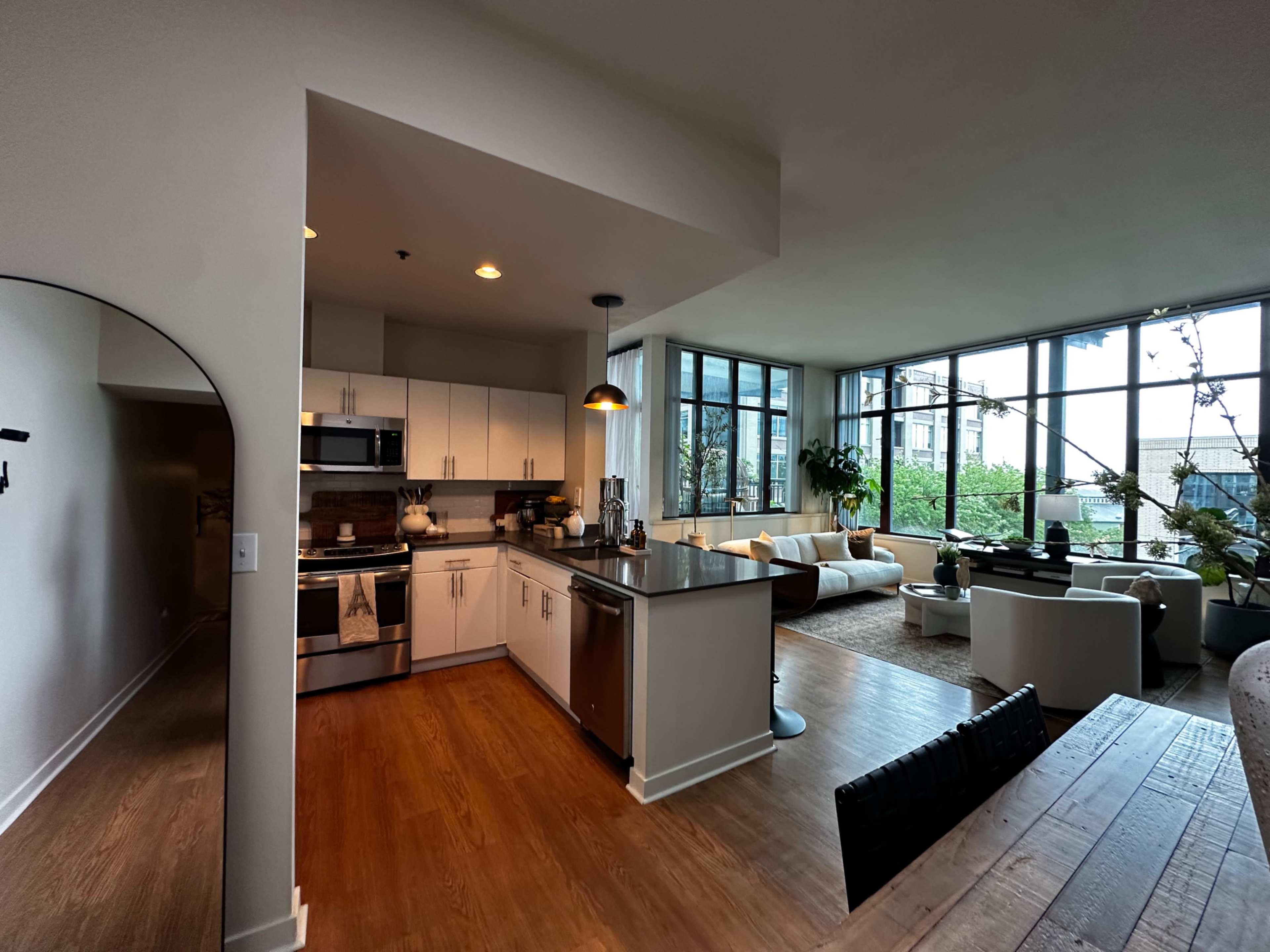 The image shows a modern open-concept living space featuring a kitchen with stainless steel appliances and a living area filled with natural light from large windows.
