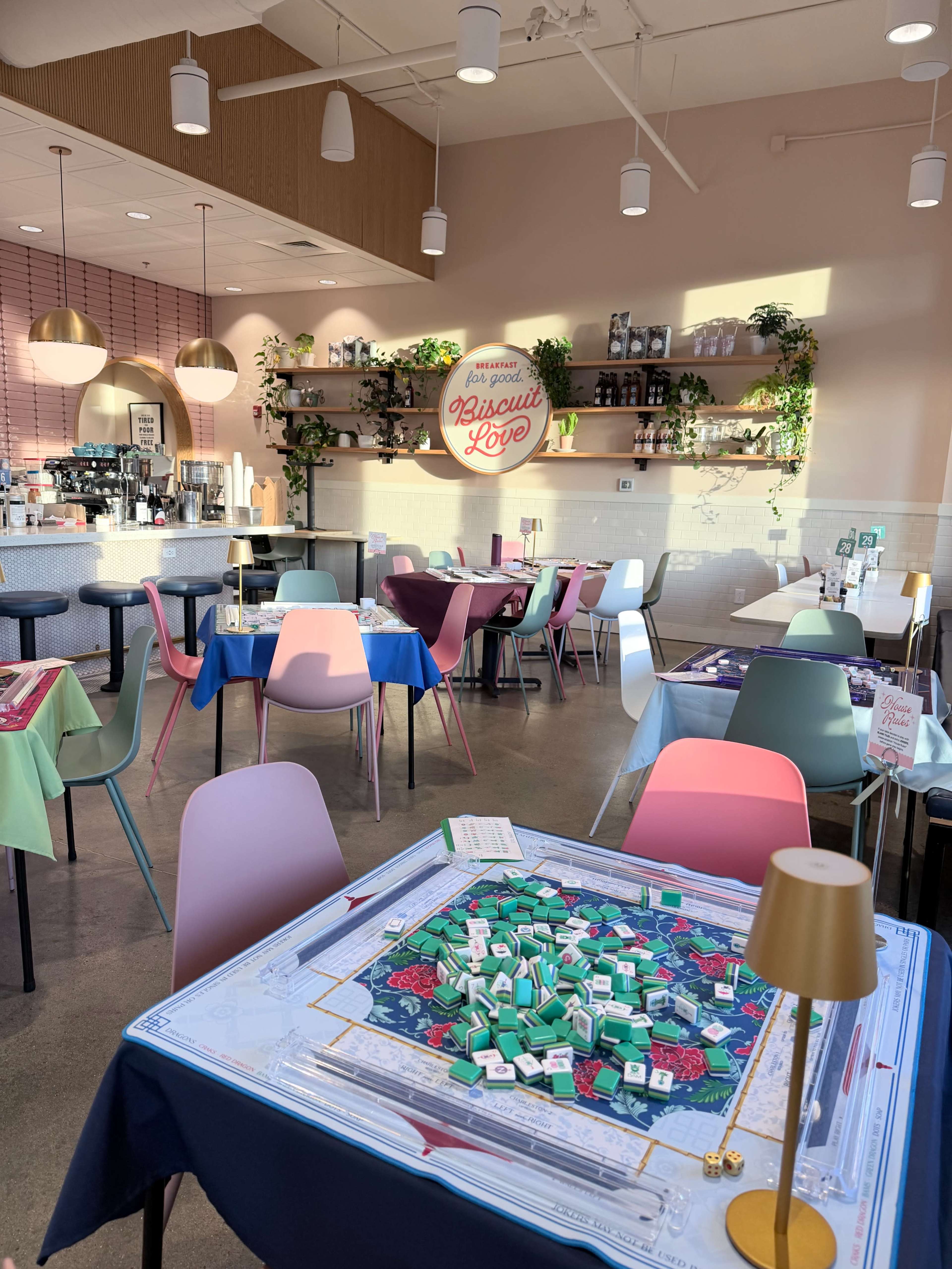 The image shows a brightly lit café with colorful chairs, a Monopoly game set up on a table, and decorative shelving in the background.