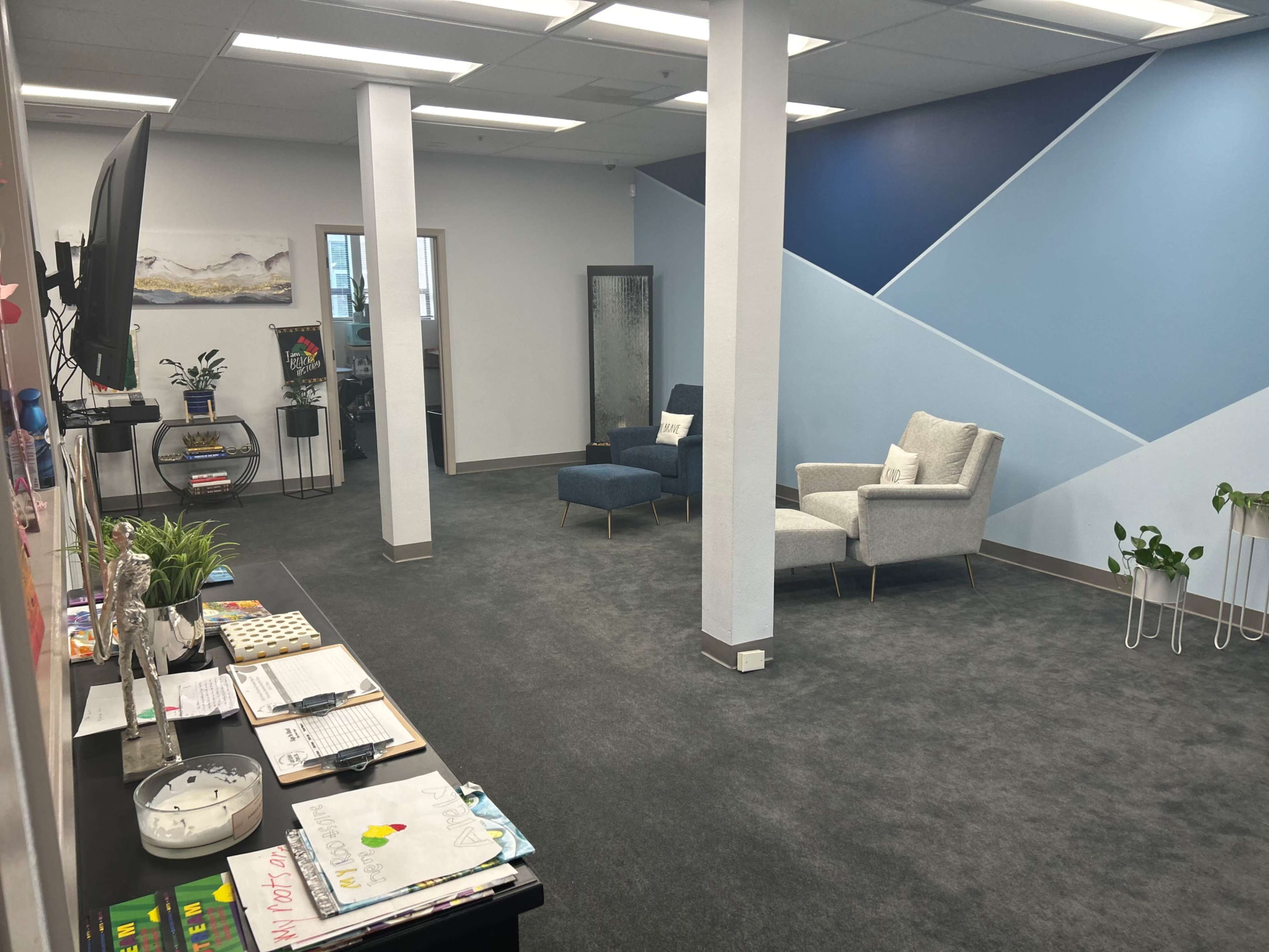 A modern office interior with a waiting area featuring two chairs, a desk with various items, and a geometric blue wall design.