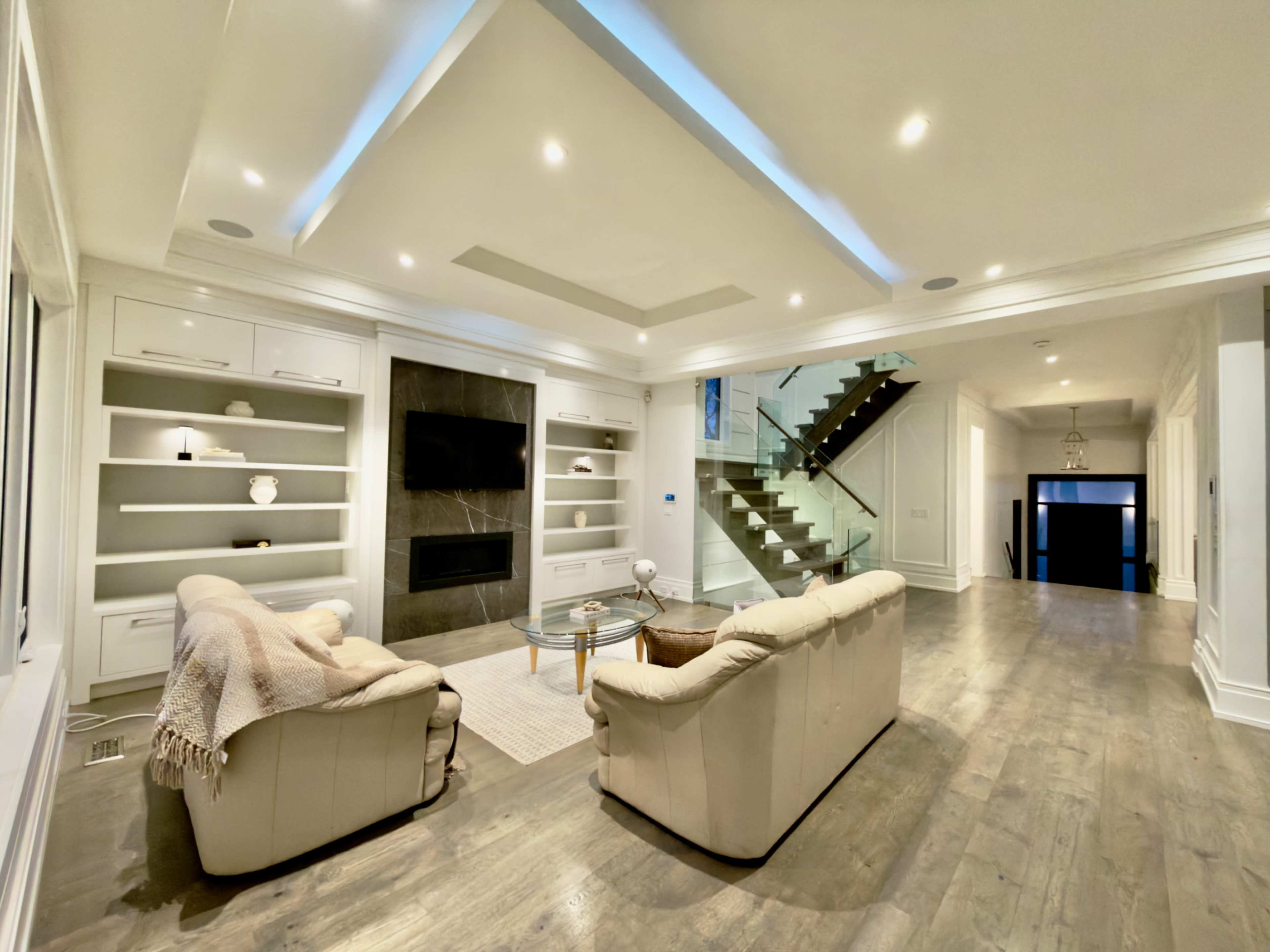 The image shows a modern living room with two light-colored sofas, a glass coffee table, built-in shelves, and a staircase in the background.
