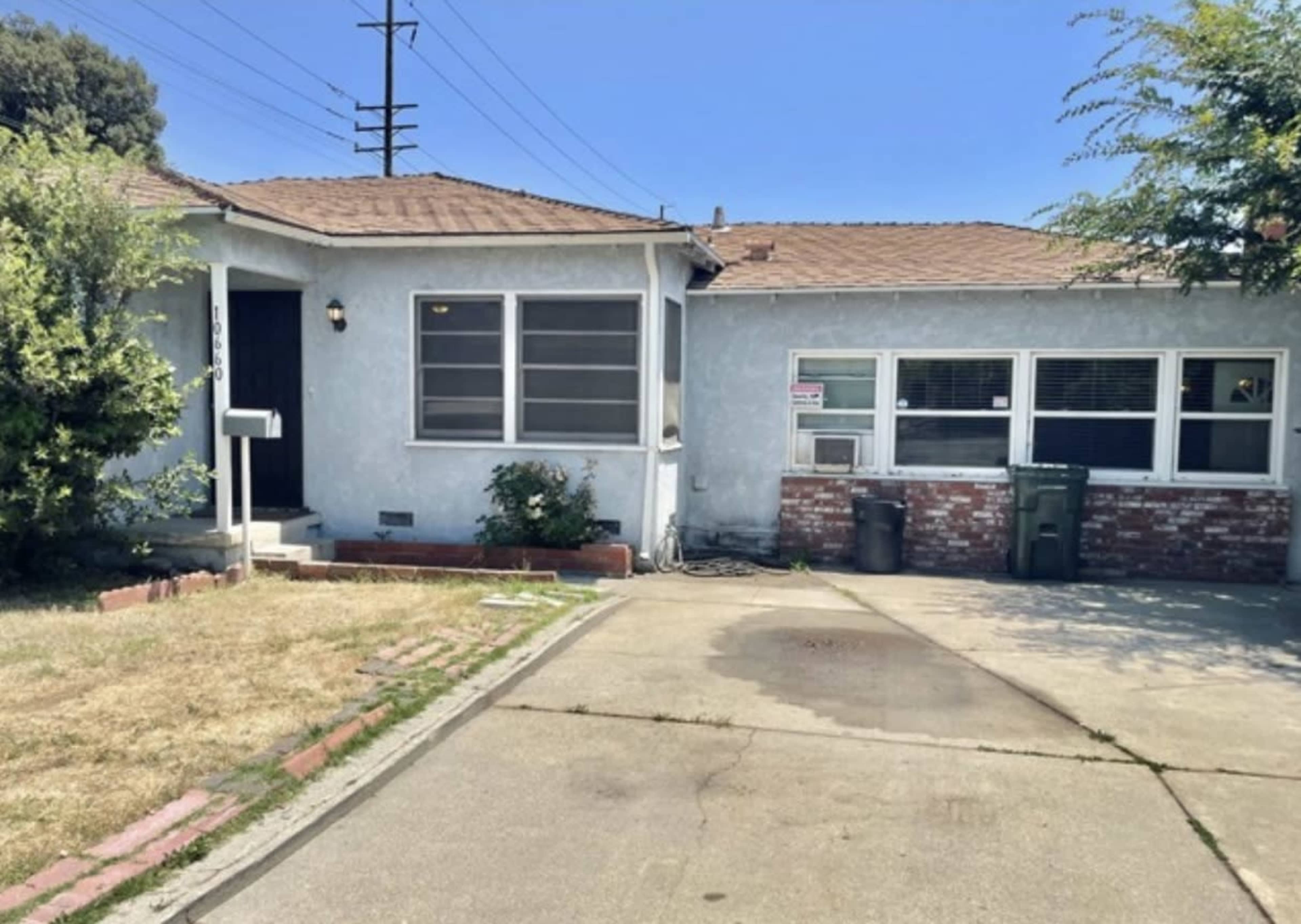The image shows a single-story house with a light blue exterior, a small front yard with patchy grass, and a paved driveway.