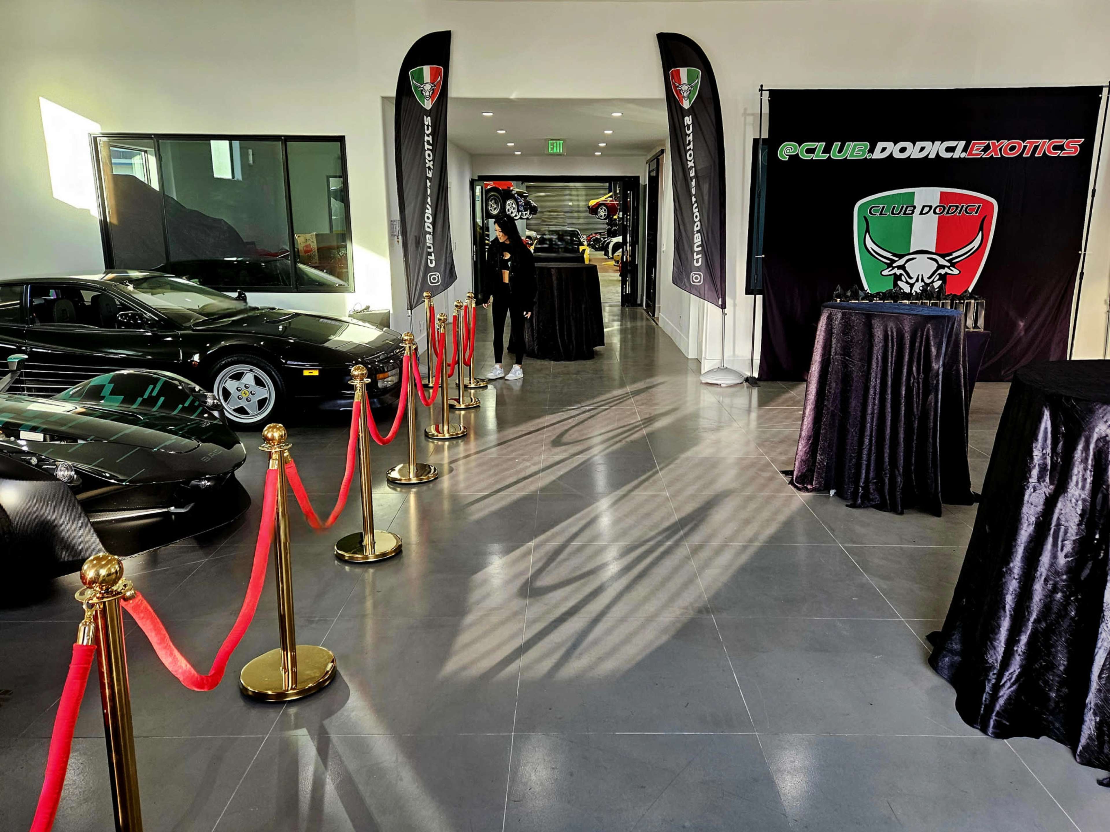The image shows a sleek car showroom with luxury vehicles displayed in the background, red ropes guiding the walkway, and black banners featuring a logo for "Club Dodici Exotics."