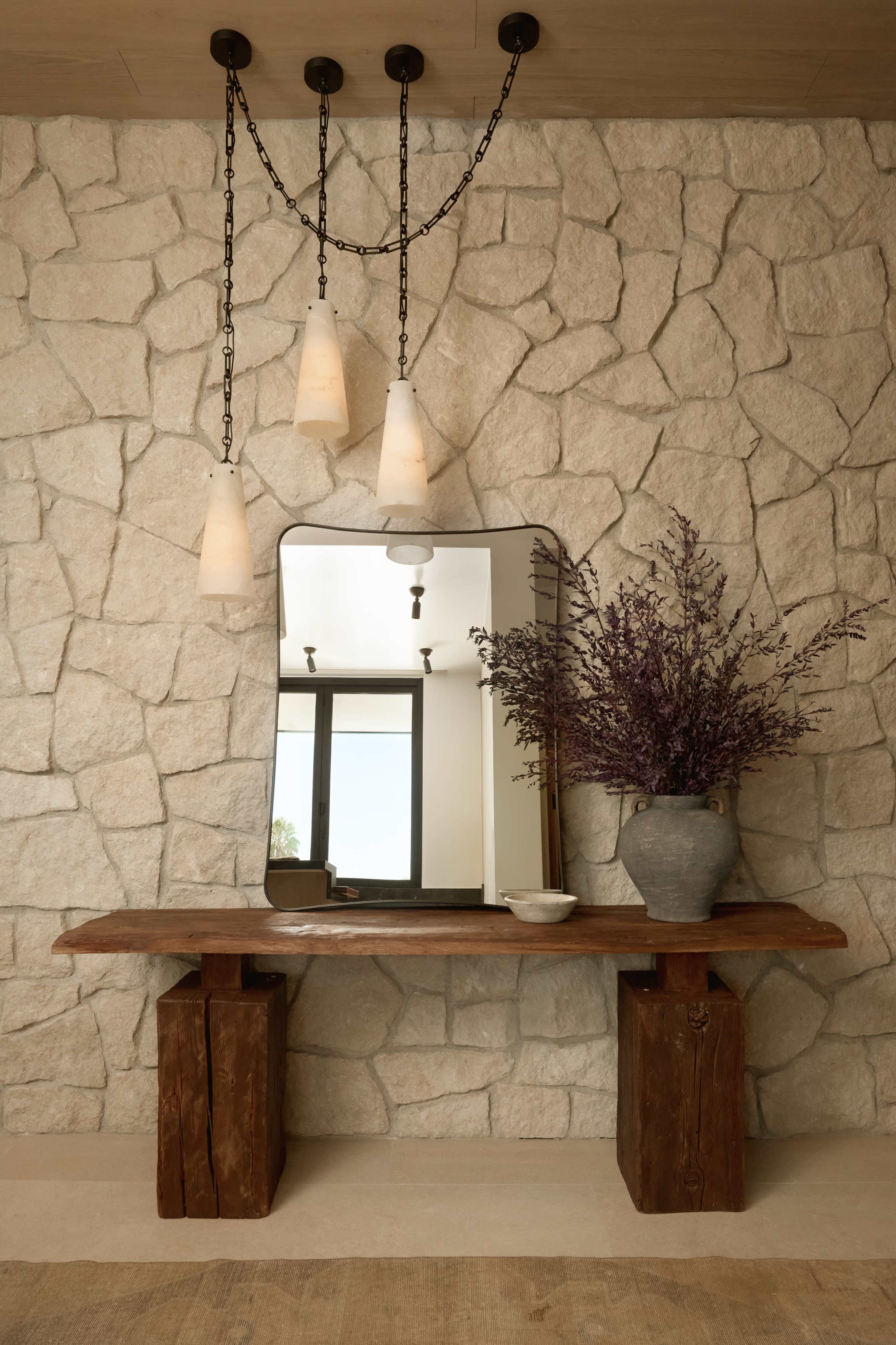 A wooden console table with a large mirror and a vase of lavender stands against a textured stone wall, illuminated by pendant lights.