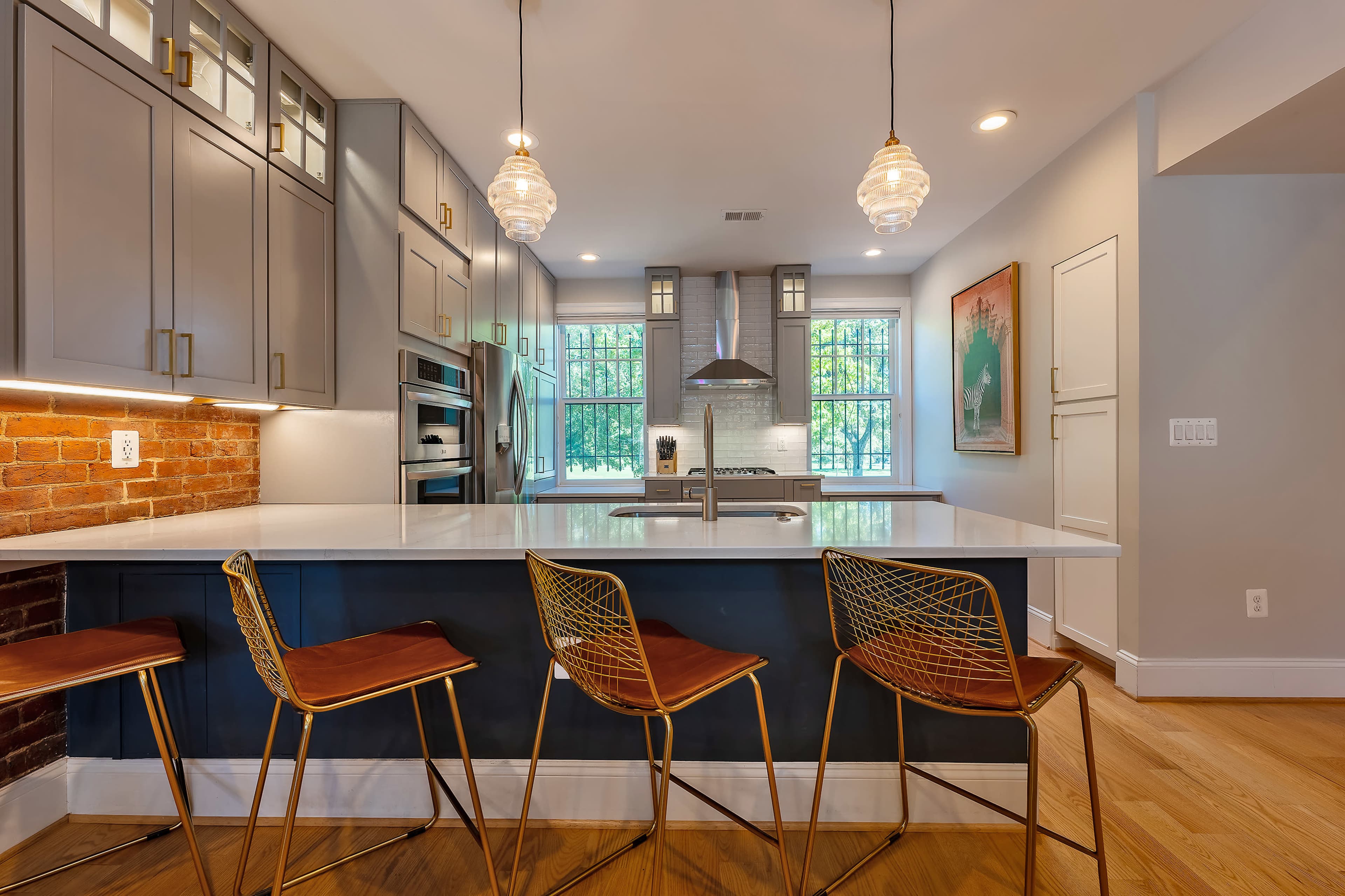 A modern kitchen features gray cabinetry, a white countertop island with gold-accented bar stools, and large windows providing natural light.