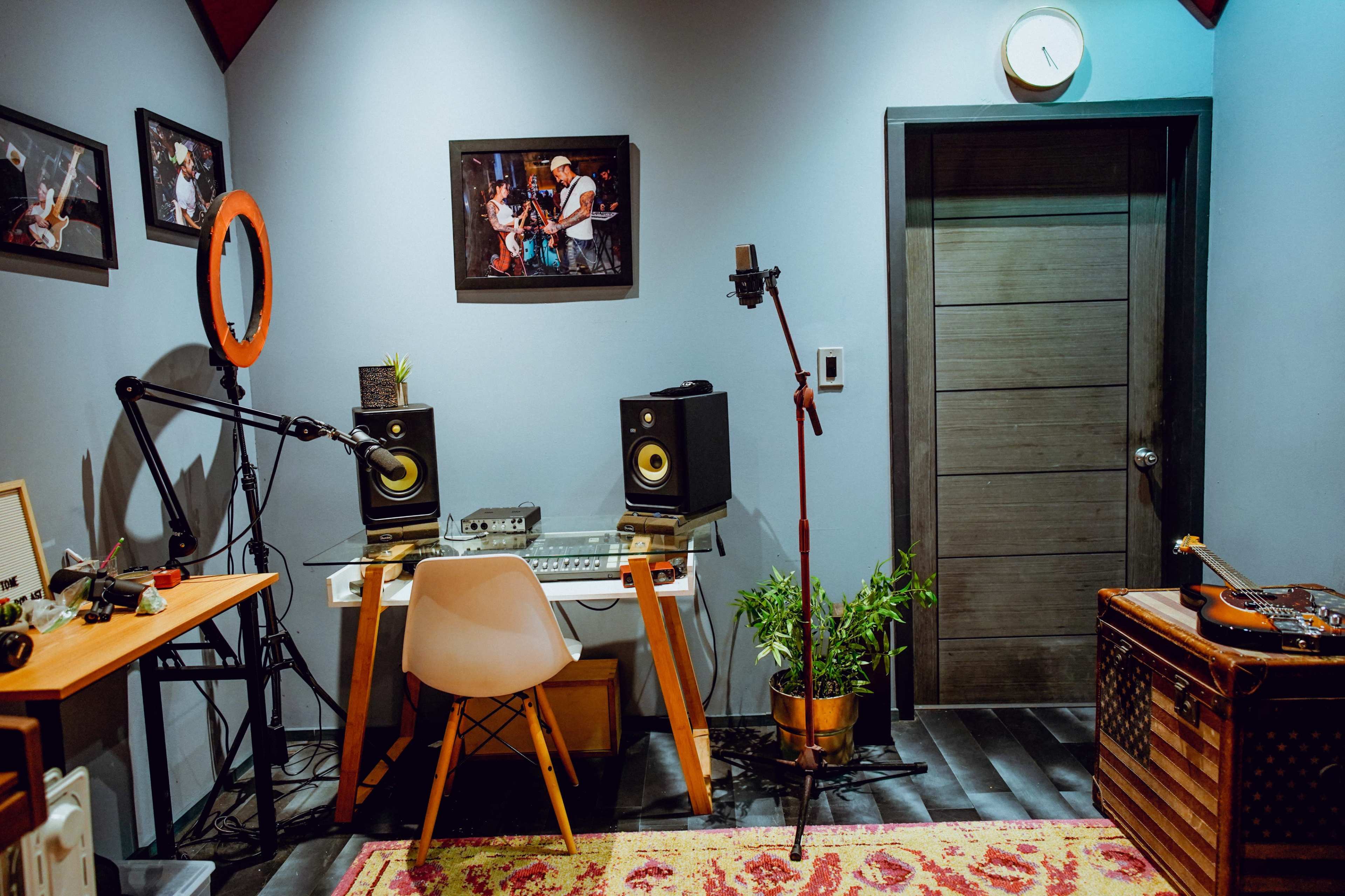 The image shows a small music studio with a desk and chair, two audio speakers, a microphone on a stand, and a potted plant, all against a wall adorned with framed photographs.