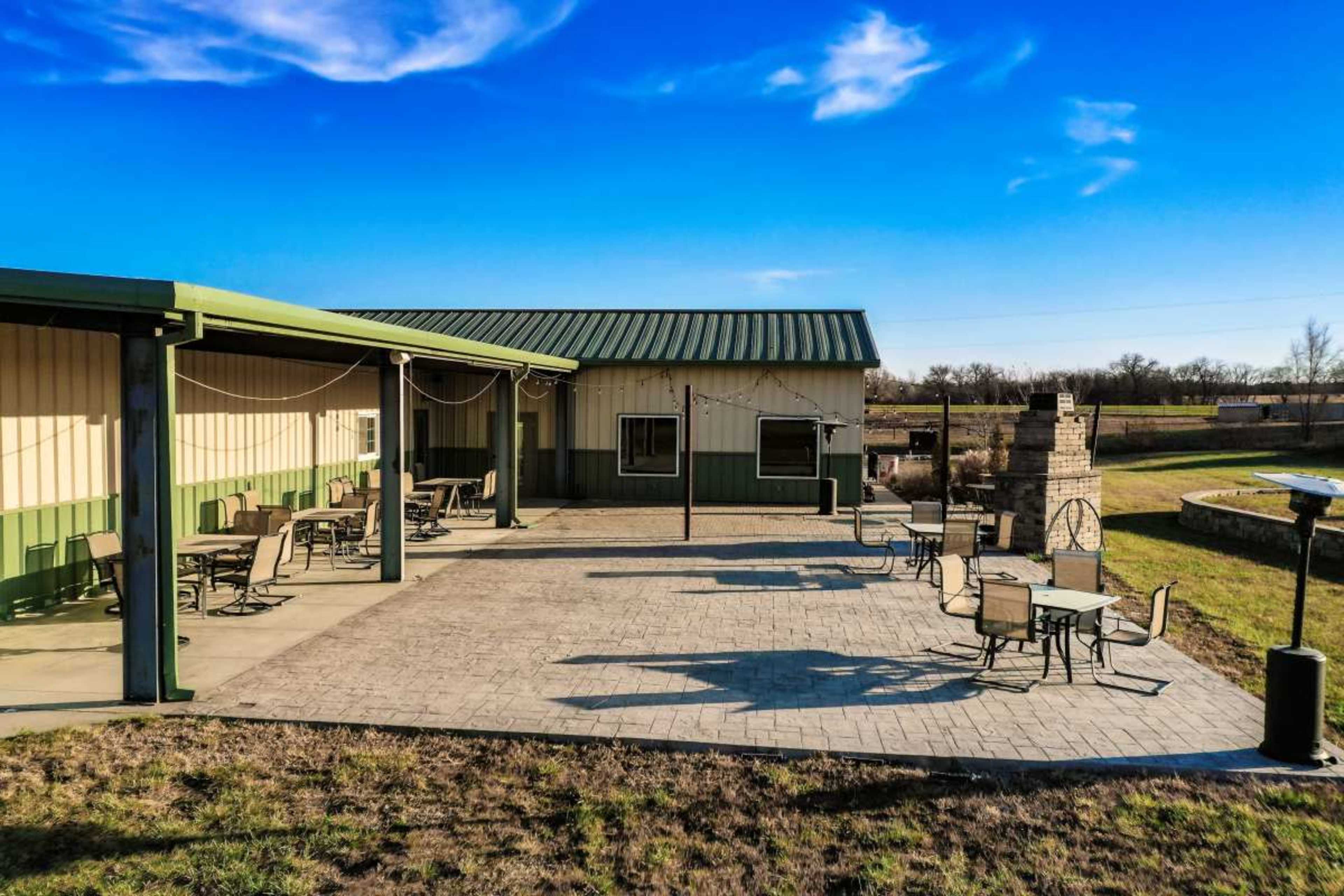 The image shows a spacious outdoor patio area with several tables and chairs, adjacent to a metal building and a stone fireplace.