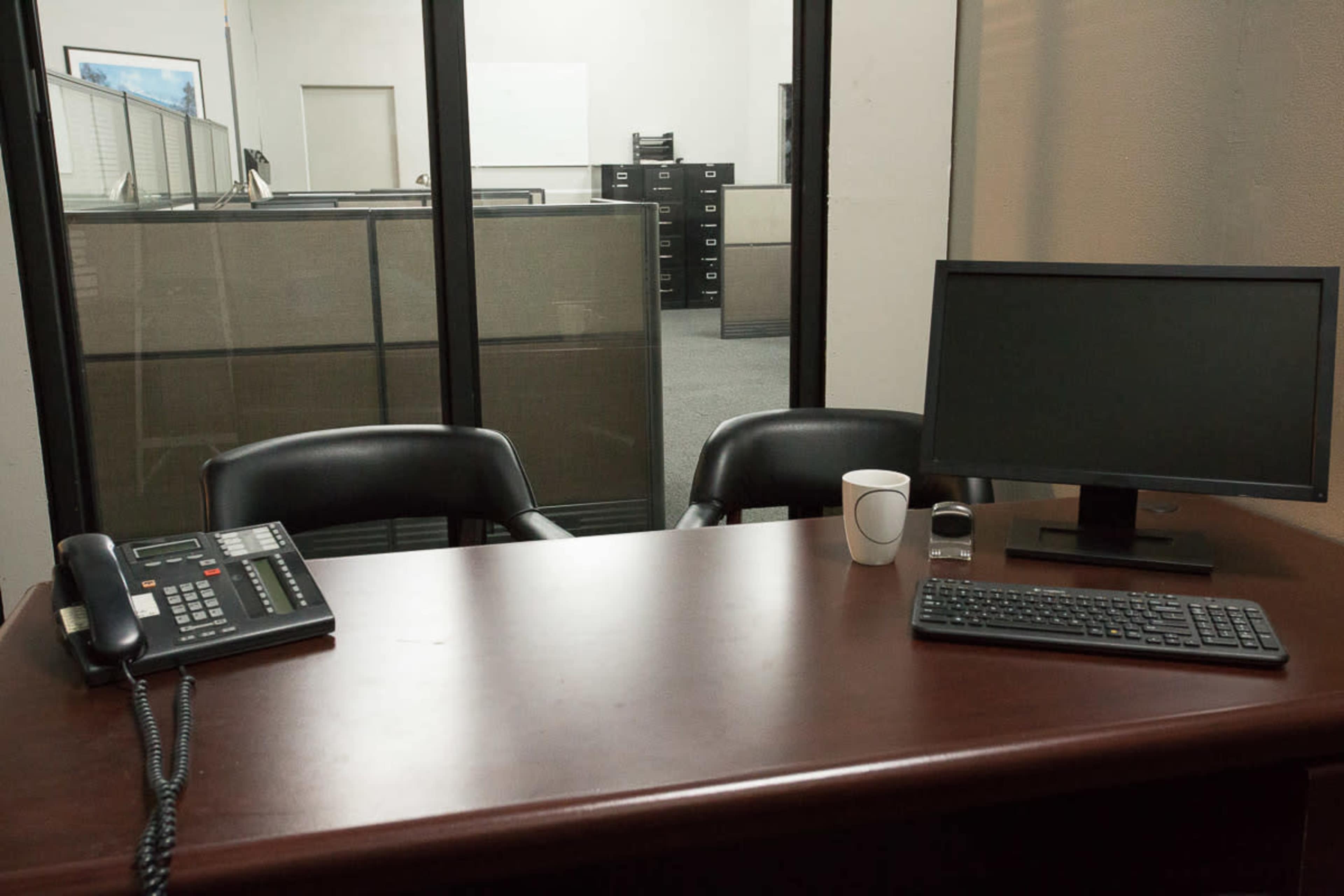 An office space features a wooden desk with a phone, monitor, keyboard, and a coffee cup, overlooking a partitioned area with filing cabinets in the background.