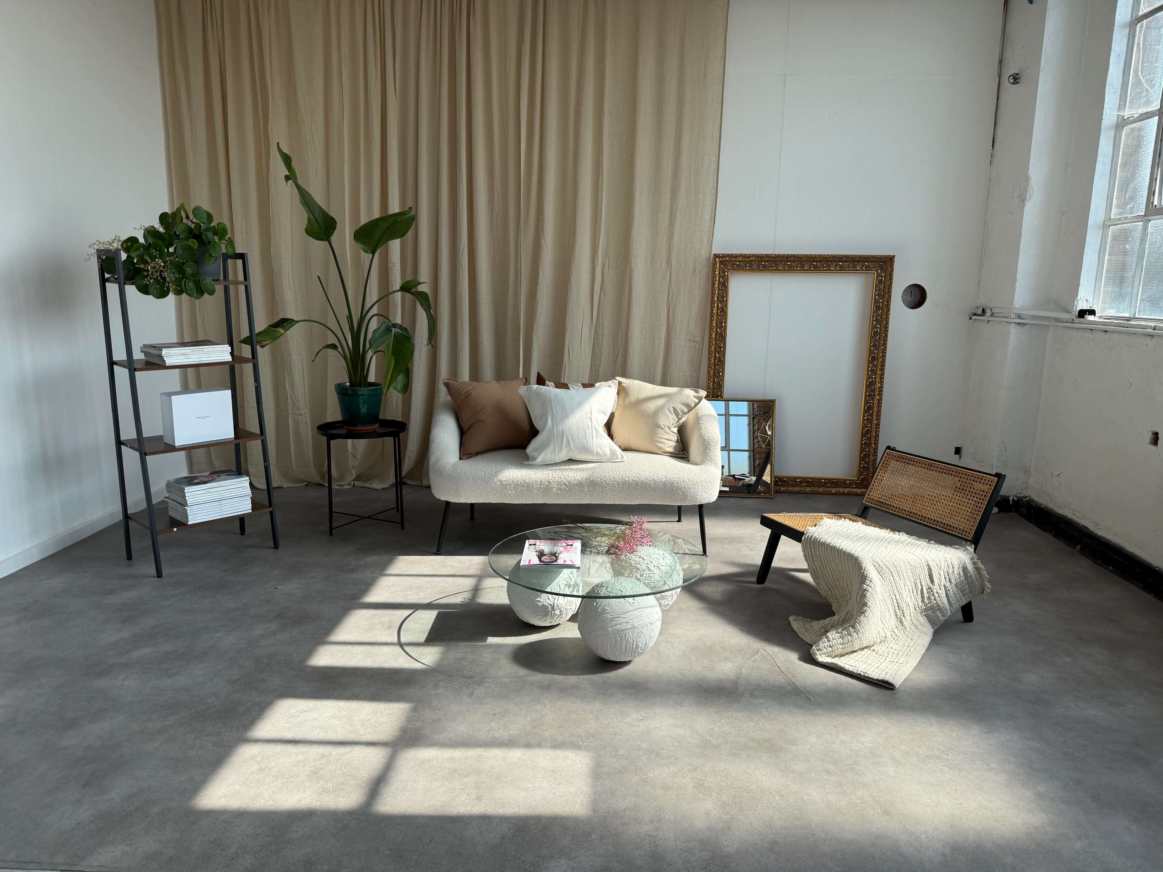 The image features a minimalist interior setting with a light-colored sofa, a plant, a glass coffee table, a stack of books, and an empty picture frame against a backdrop of beige curtains.