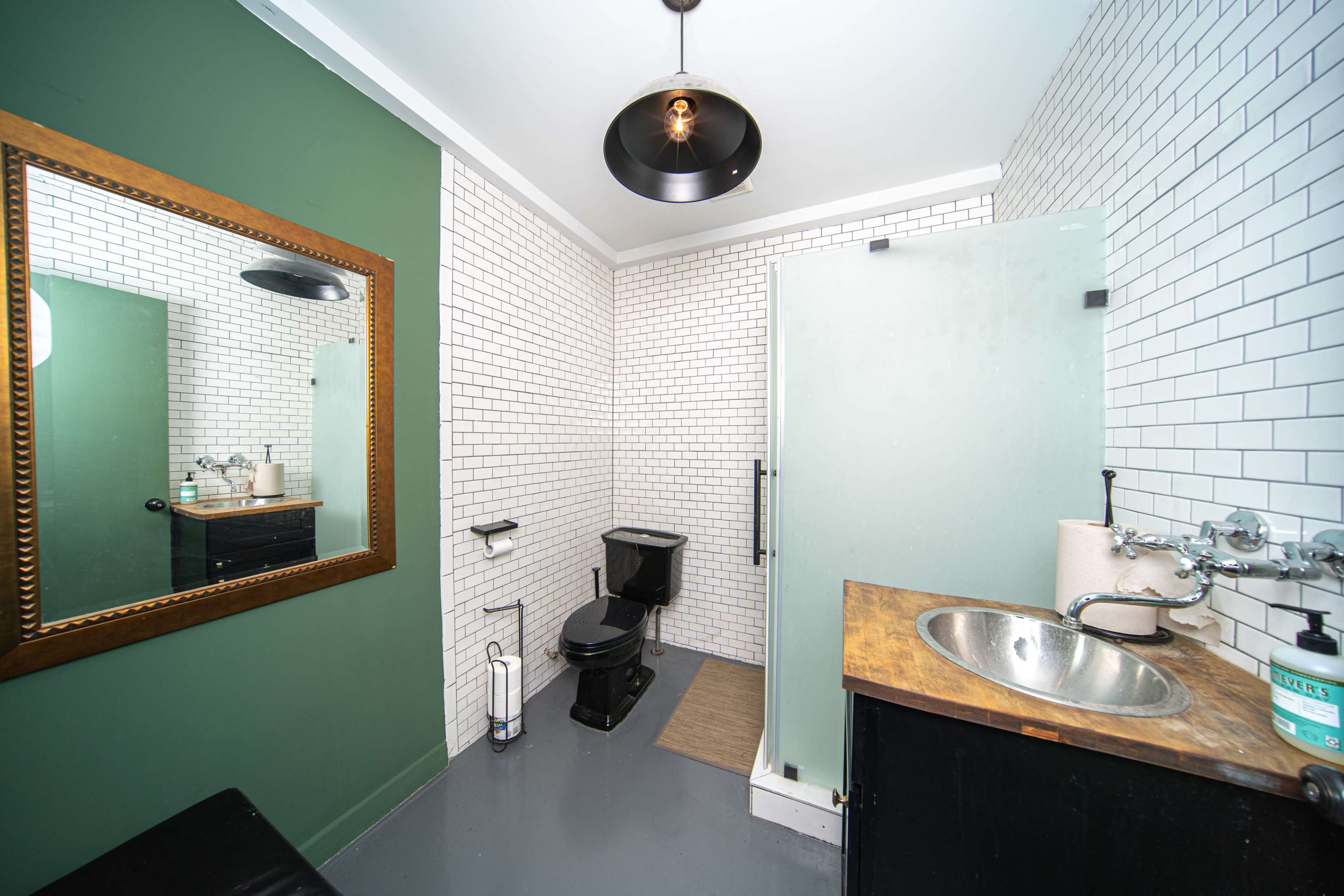 The image shows a modern bathroom with white tiled walls, a wooden sink, a black toilet, and a large mirror.