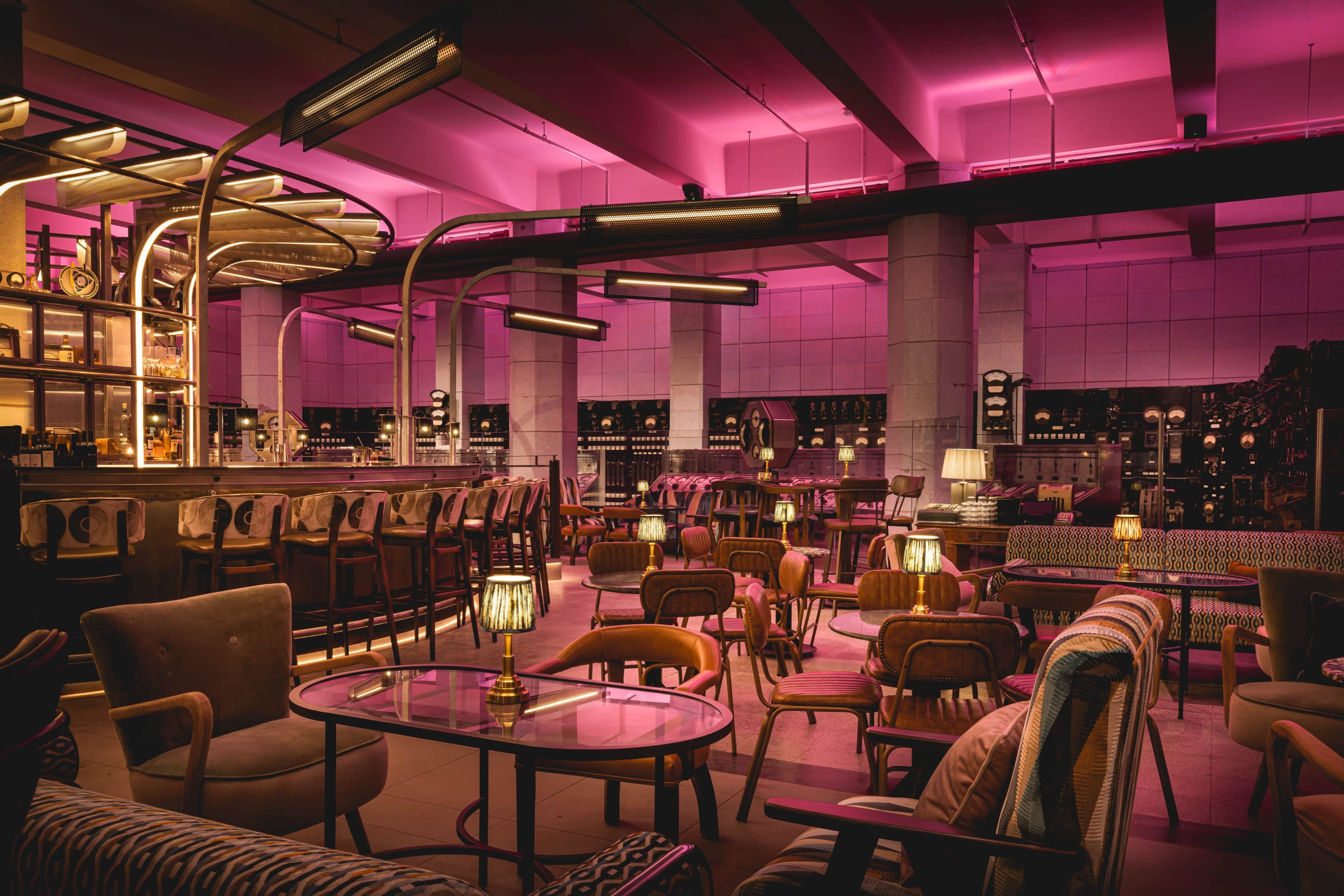 A dimly lit bar with pink lighting, featuring a variety of seating options and a large bar area filled with bottles.