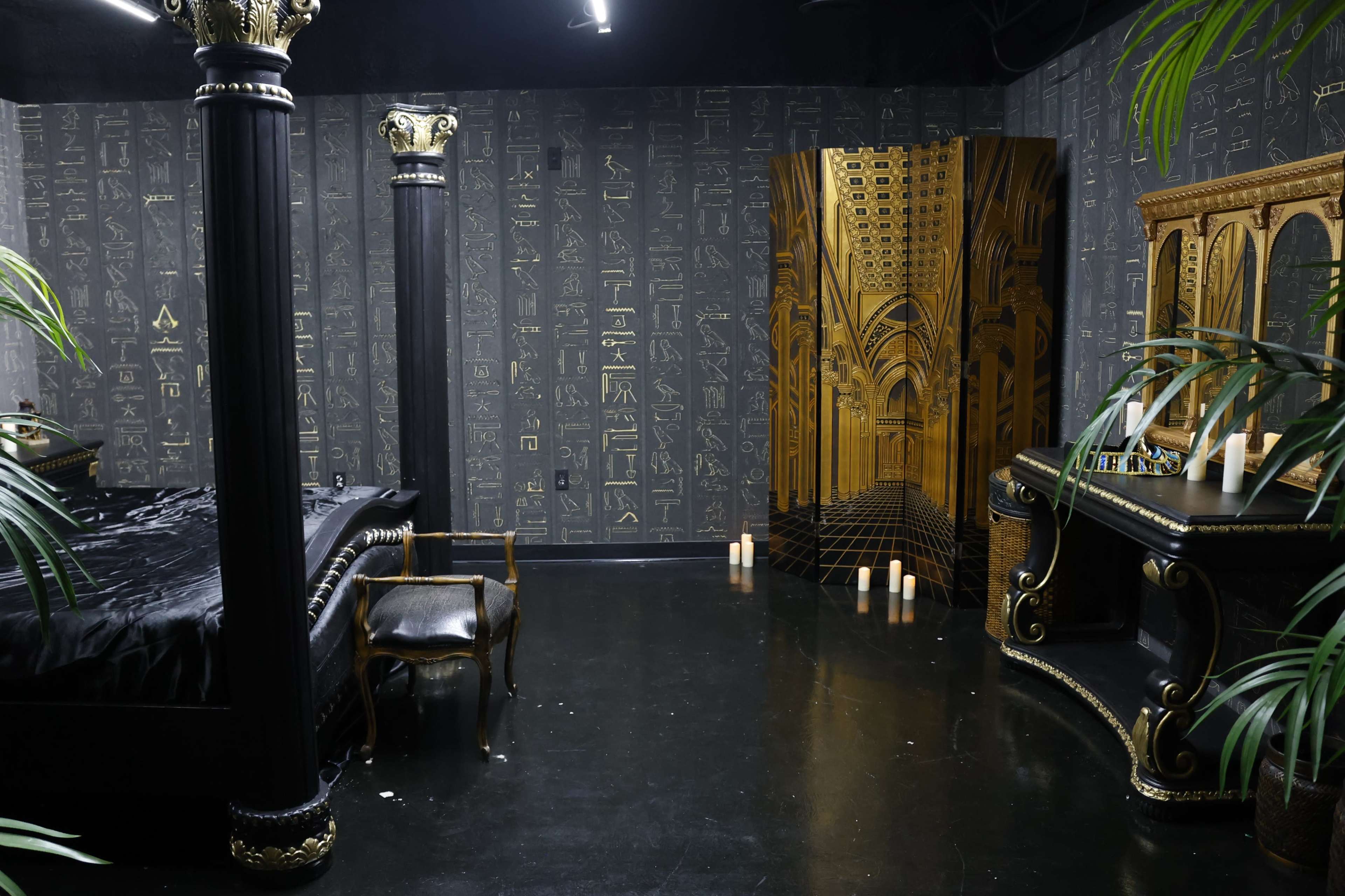 A dimly lit room featuring a black bed, ornate pillars, and decorative gold accents on the furniture, with candles positioned on the floor.