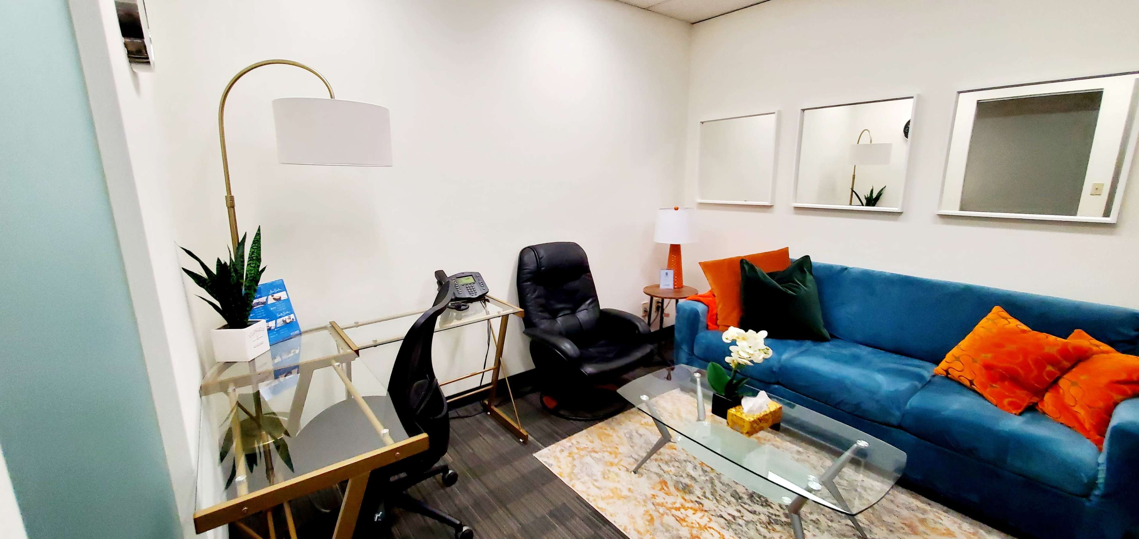 A small office space with a blue couch, a glass coffee table, a desk with a lamp, and a black chair.
