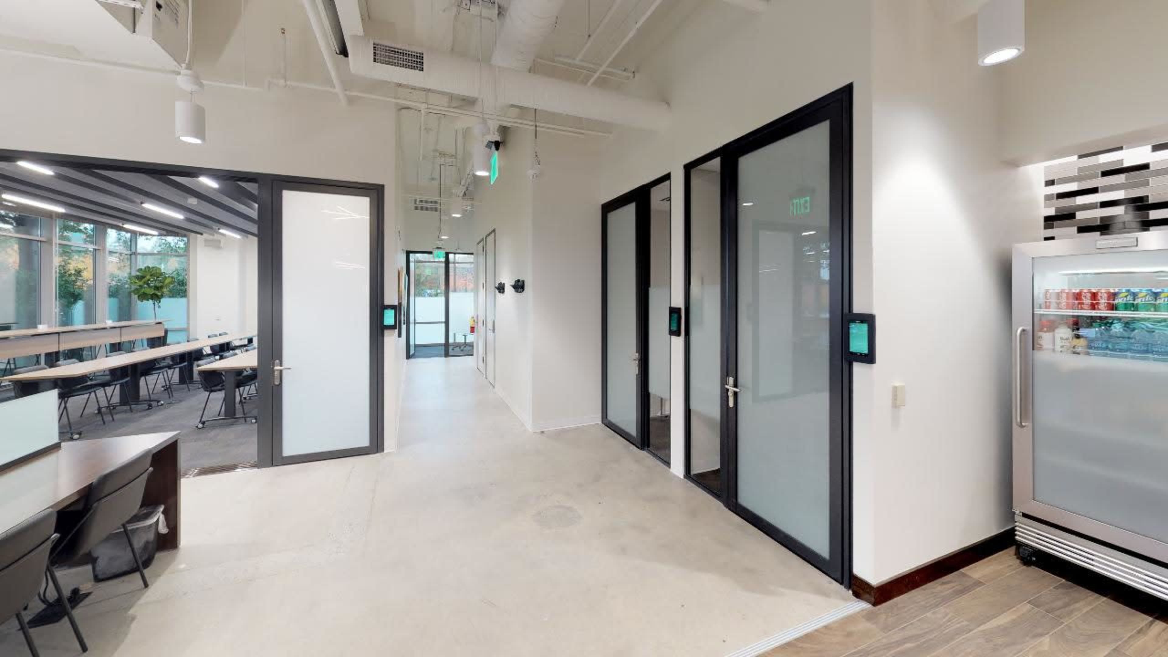 A modern office space with glass-encased meeting rooms, a refrigerator against one wall, and a communal seating area.