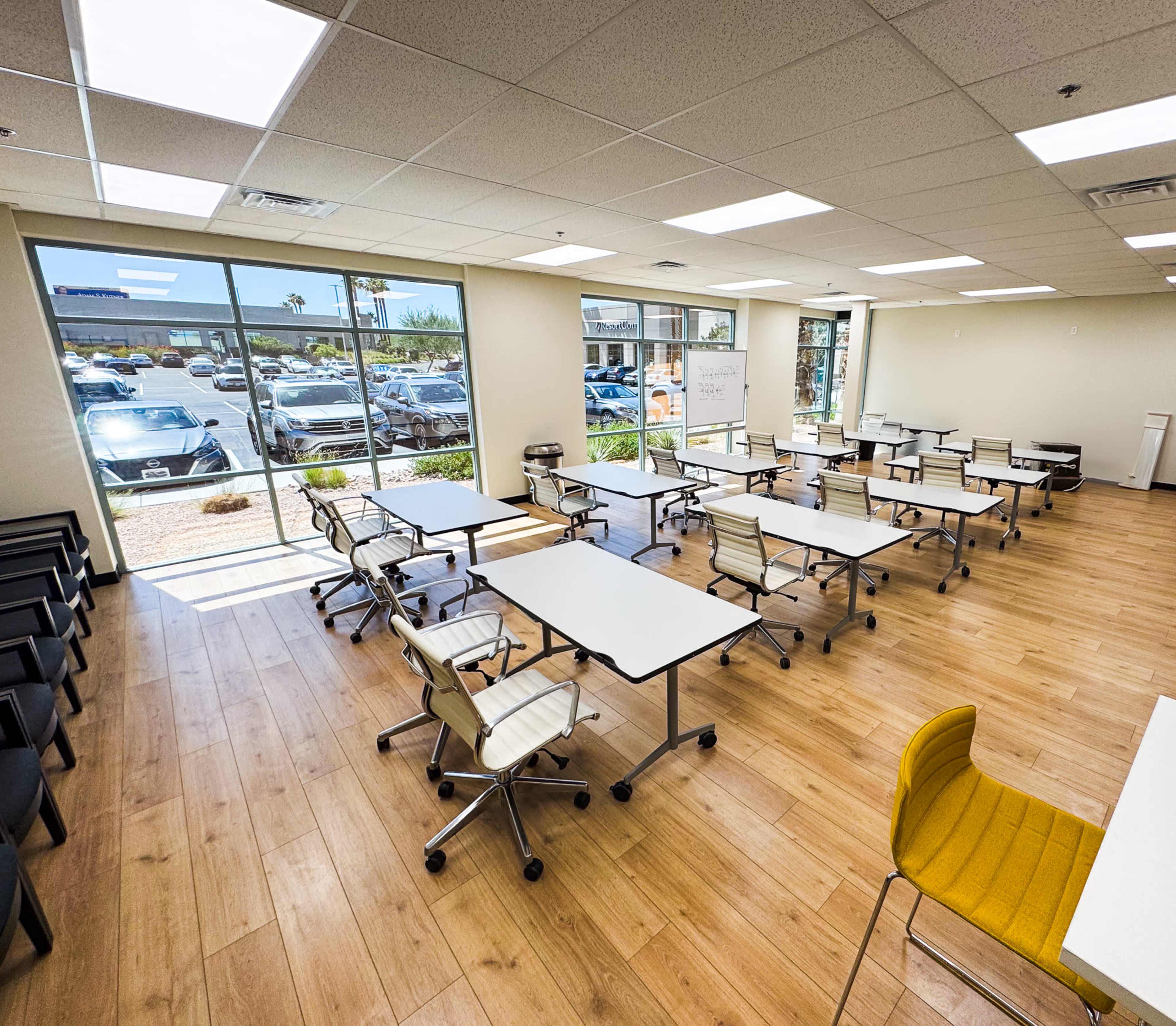 The image shows a modern classroom setup with white tables and rolling chairs arranged in a spacious room featuring large windows and wooden flooring.