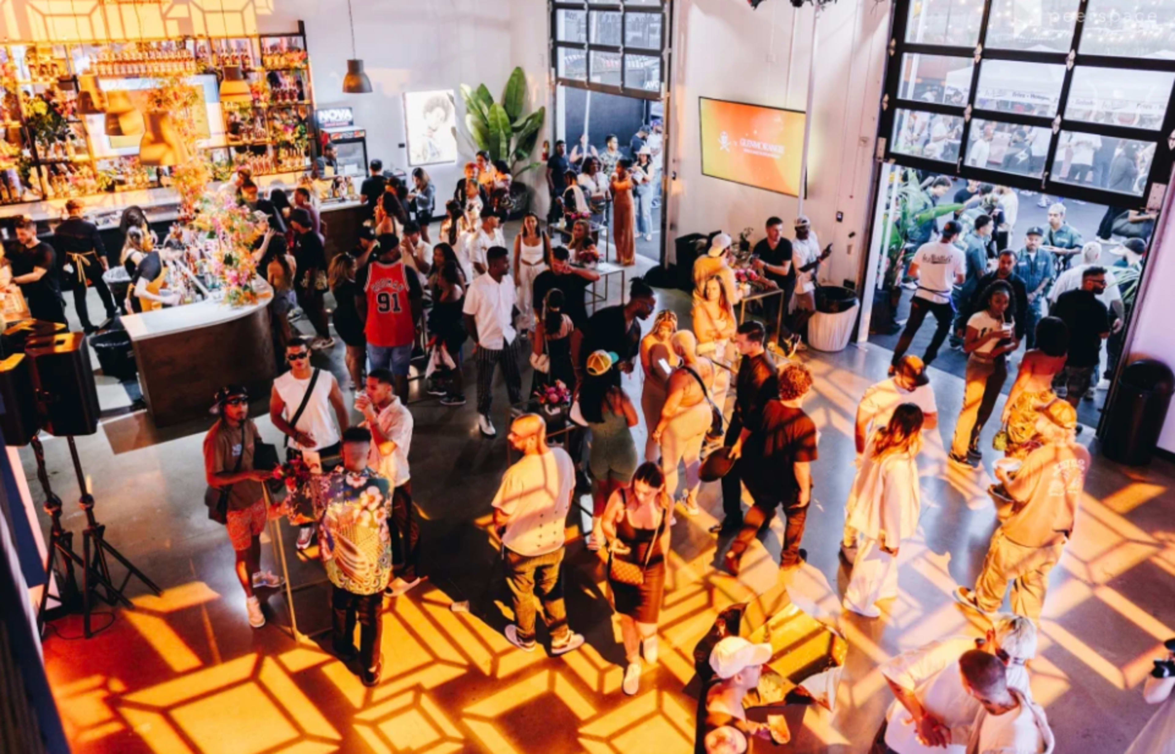 A large crowd gathers in a vibrant event space featuring a bar and a dance floor, illuminated by bright patterns from overhead lighting.