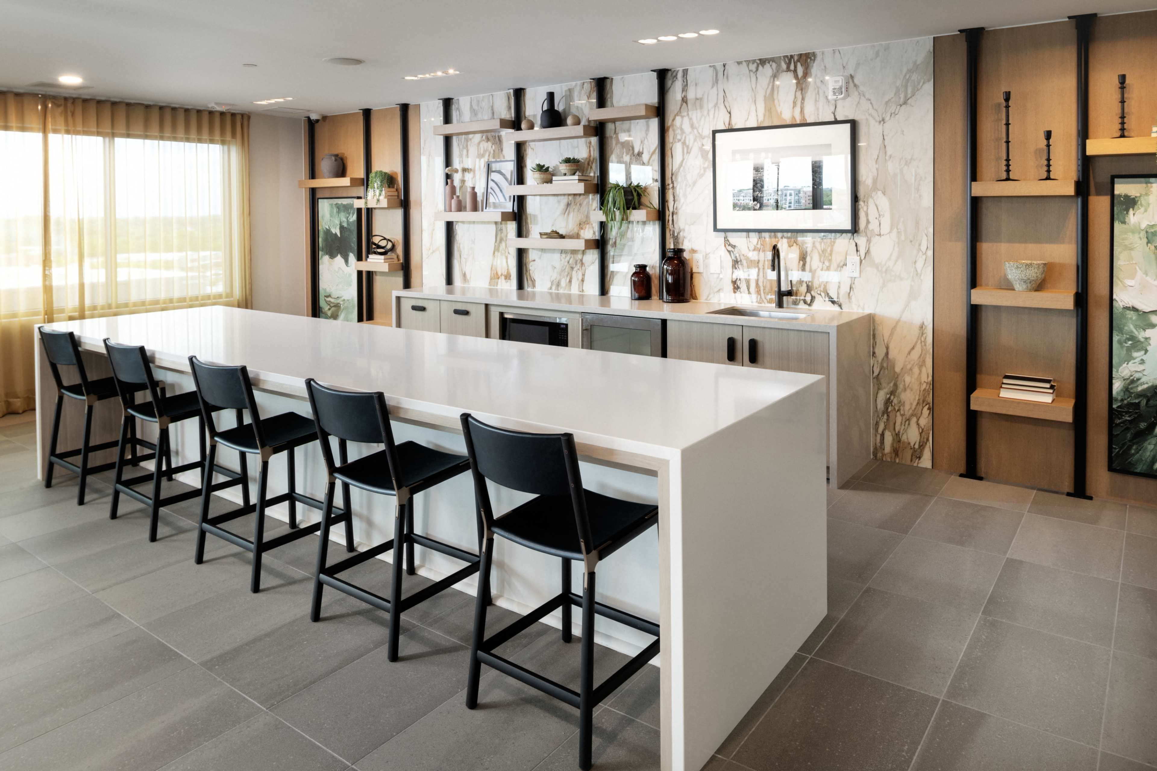 The image shows a modern kitchen with a white island countertop, black bar stools, and decorative shelves featuring plants and artwork.