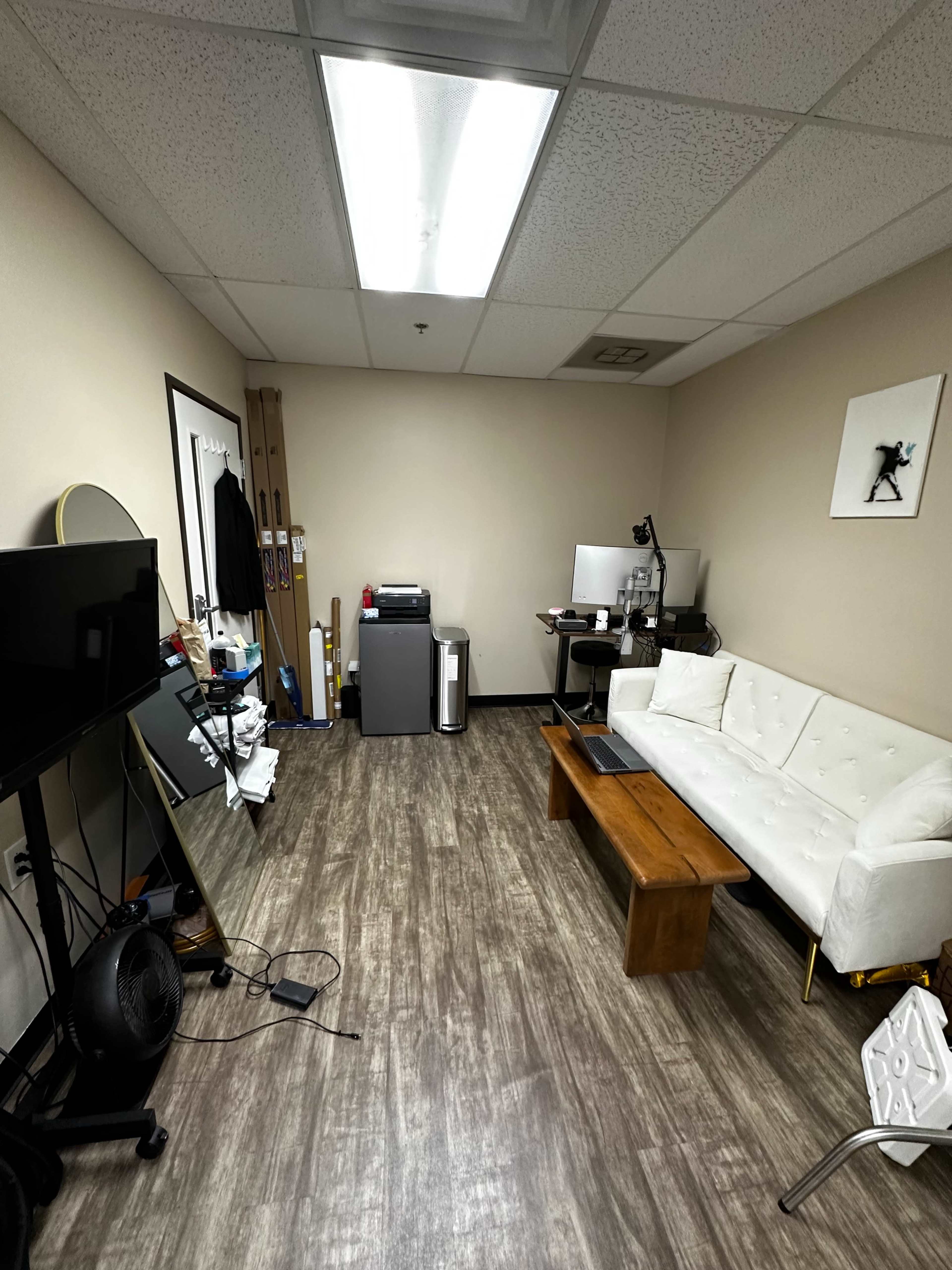An office space with a white sofa, a wooden coffee table, a television, and a computer setup against a light-colored wall.