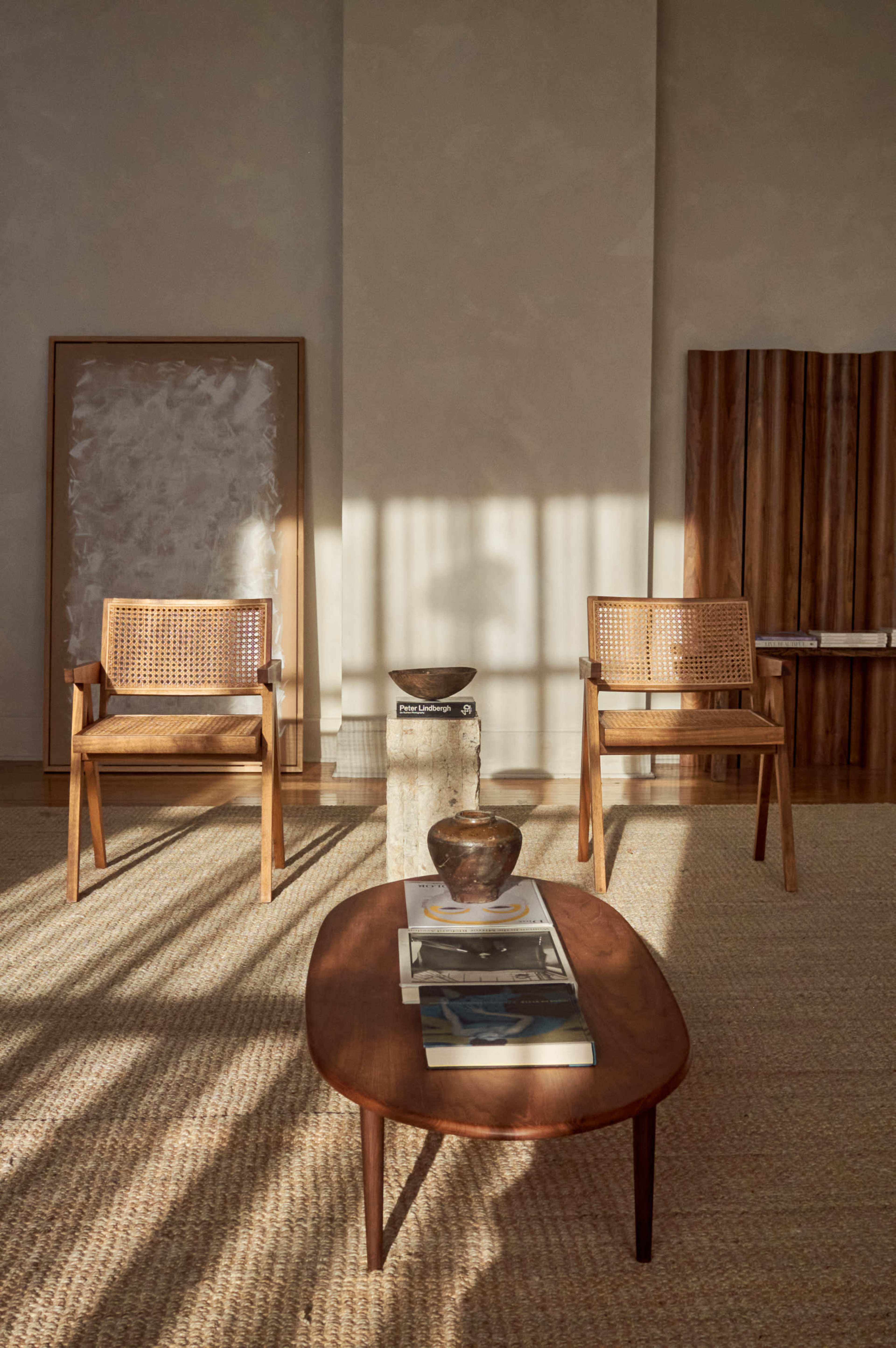 The image shows a minimalist interior with two woven chairs, a low wooden coffee table stacked with books, and a decorative bowl, all illuminated by soft, natural light.