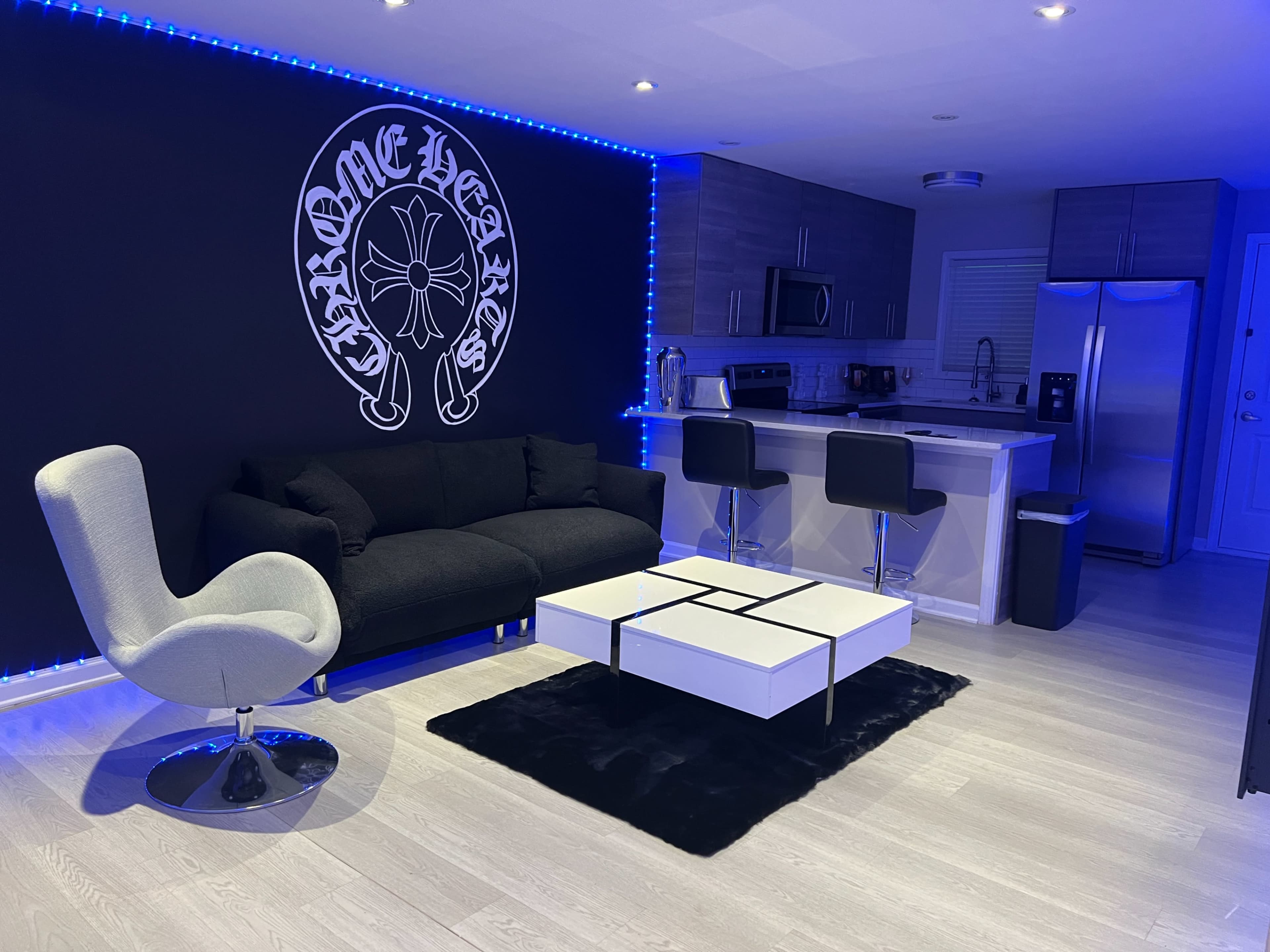 The image shows a contemporary living space featuring a black sofa, a modern coffee table, and a kitchen area with stainless steel appliances, all illuminated by blue LED lights.