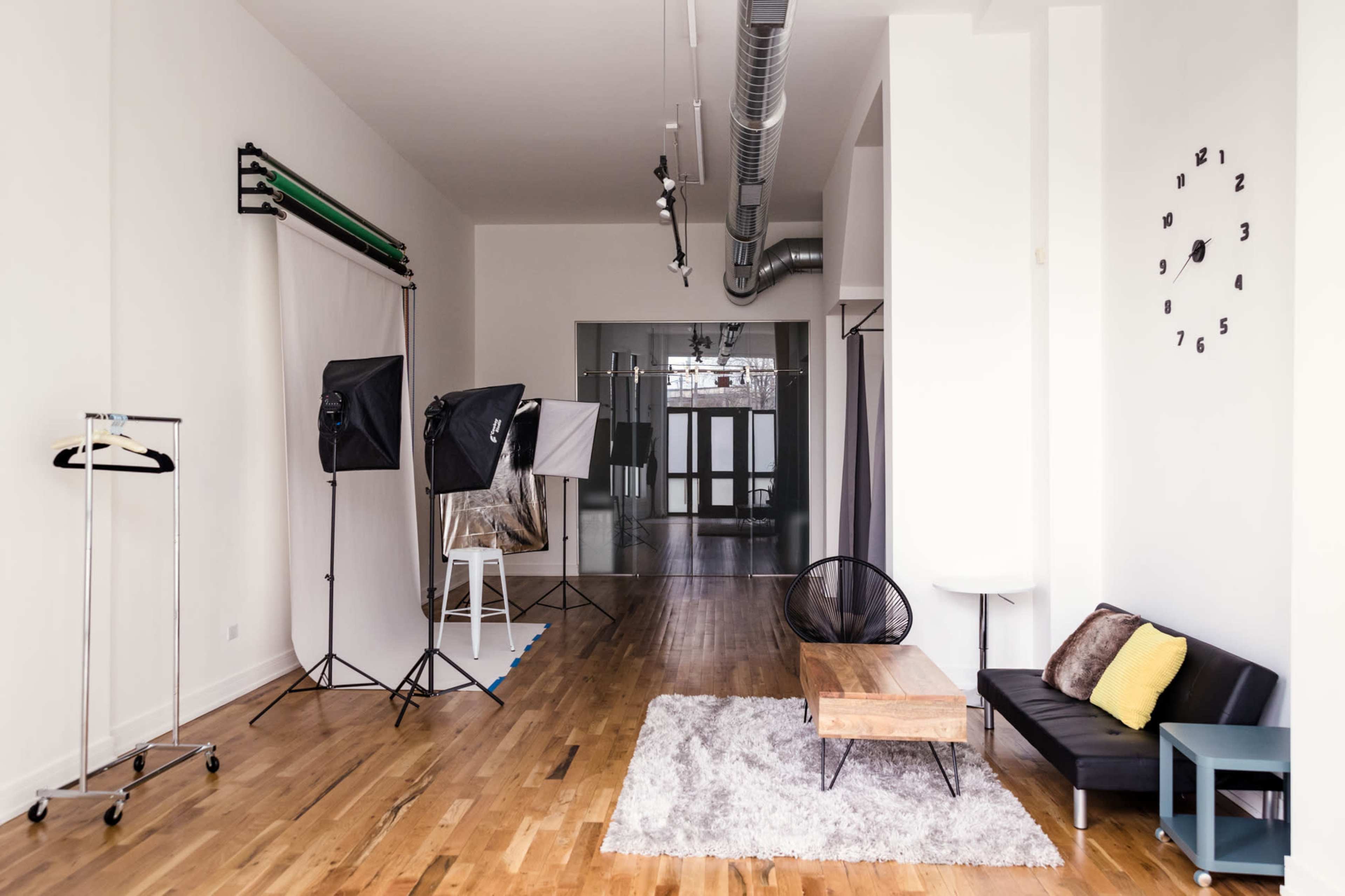 A spacious, well-lit photo studio with backdrop equipment, lighting setups, and a seating area featuring a black sofa and a wooden coffee table on a rug.