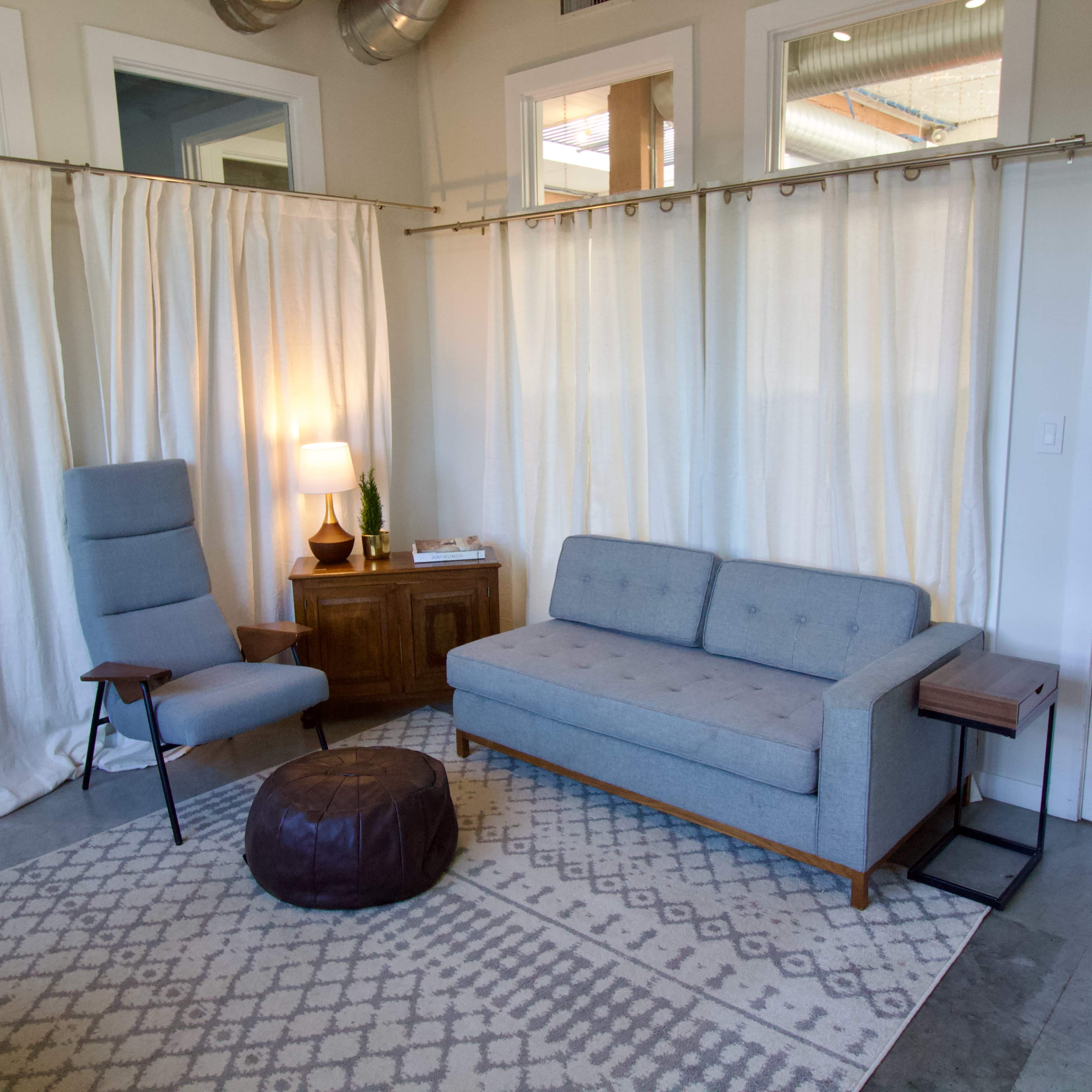 The image shows a modern living space with a gray sofa, a light gray armchair, a wooden side table, and a brown pouf, all set against a backdrop of white curtains and a patterned rug on a concrete floor.