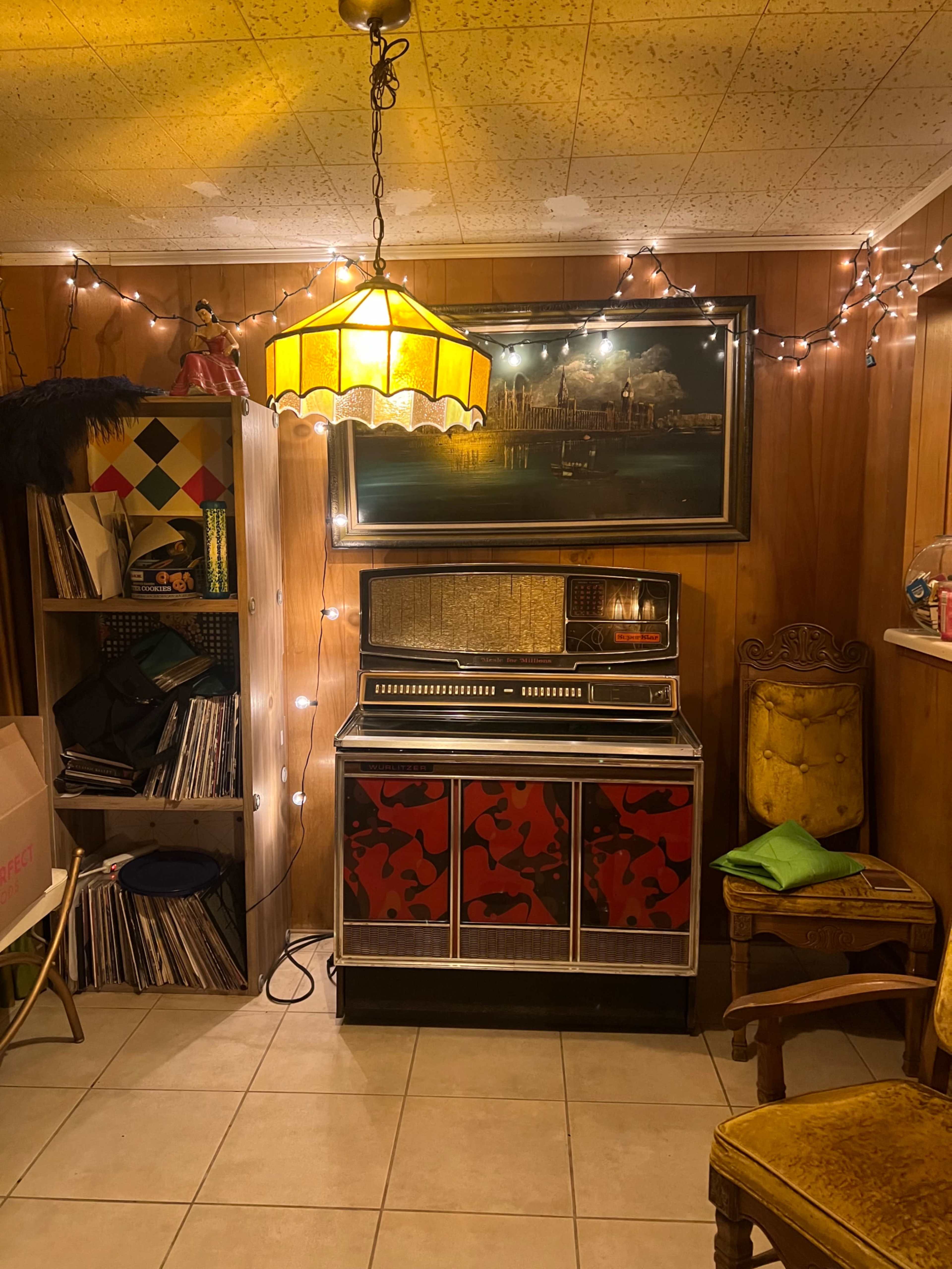 The image depicts a retro basement space featuring a vintage jukebox, a shelf of vinyl records, a decorative lamp, and fairy lights illuminating the wooden paneling.