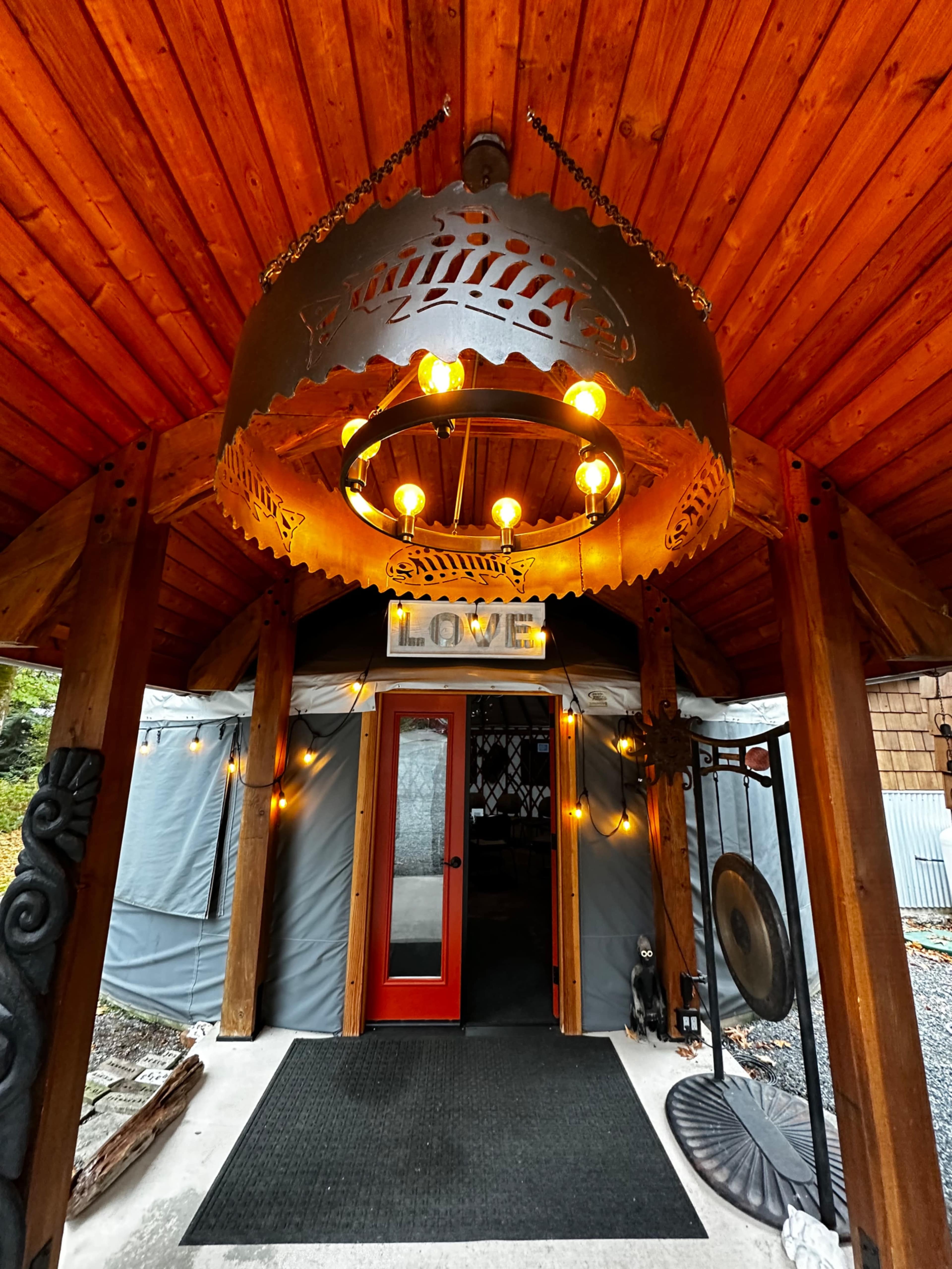 The entrance of a rustic structure features a large metal chandelier and a red door, with "LOVE" displayed above surrounded by string lights.