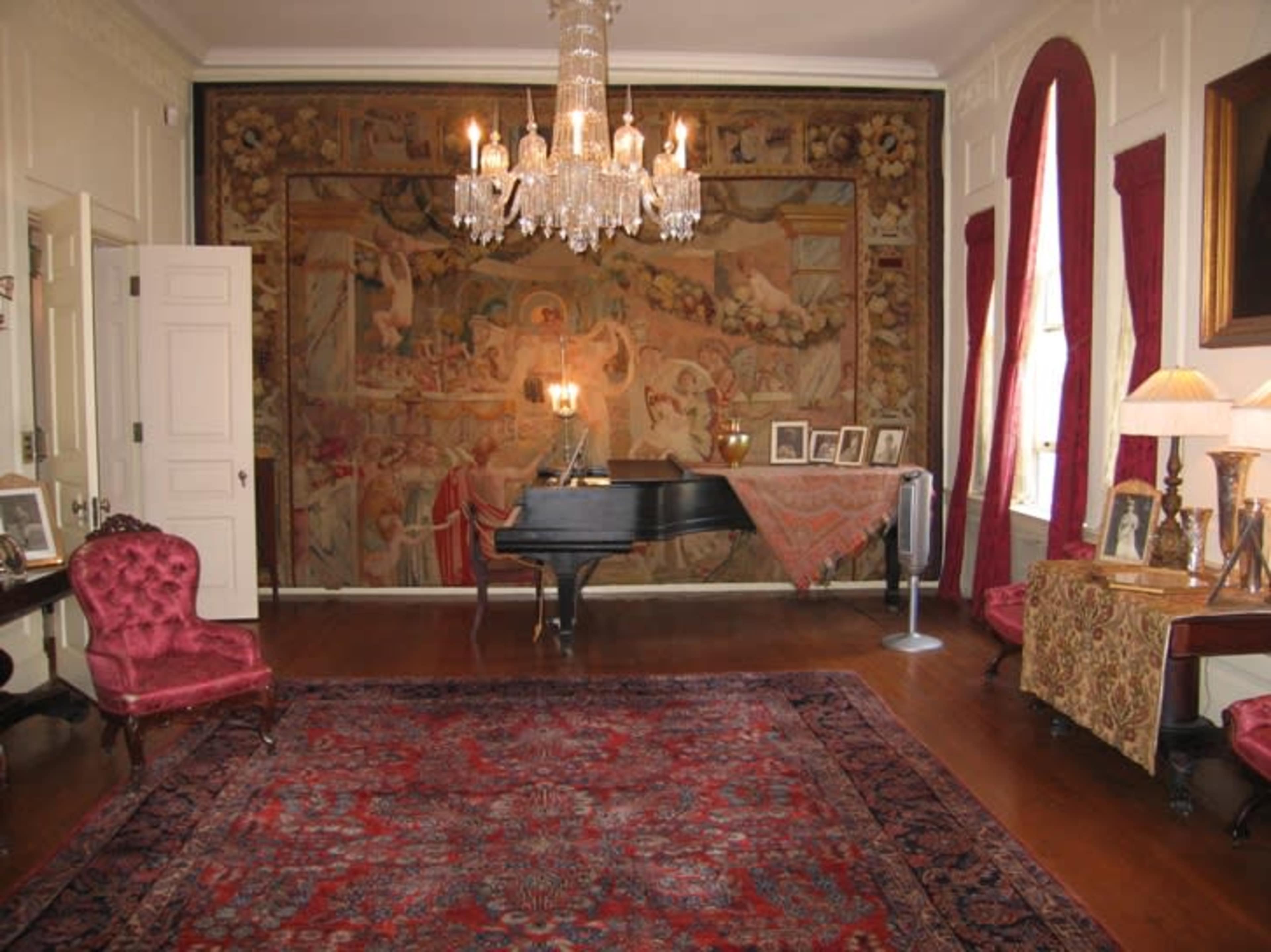 A grand room features a black grand piano, a large ornate chandelier, and a wall covered with an intricate tapestry.