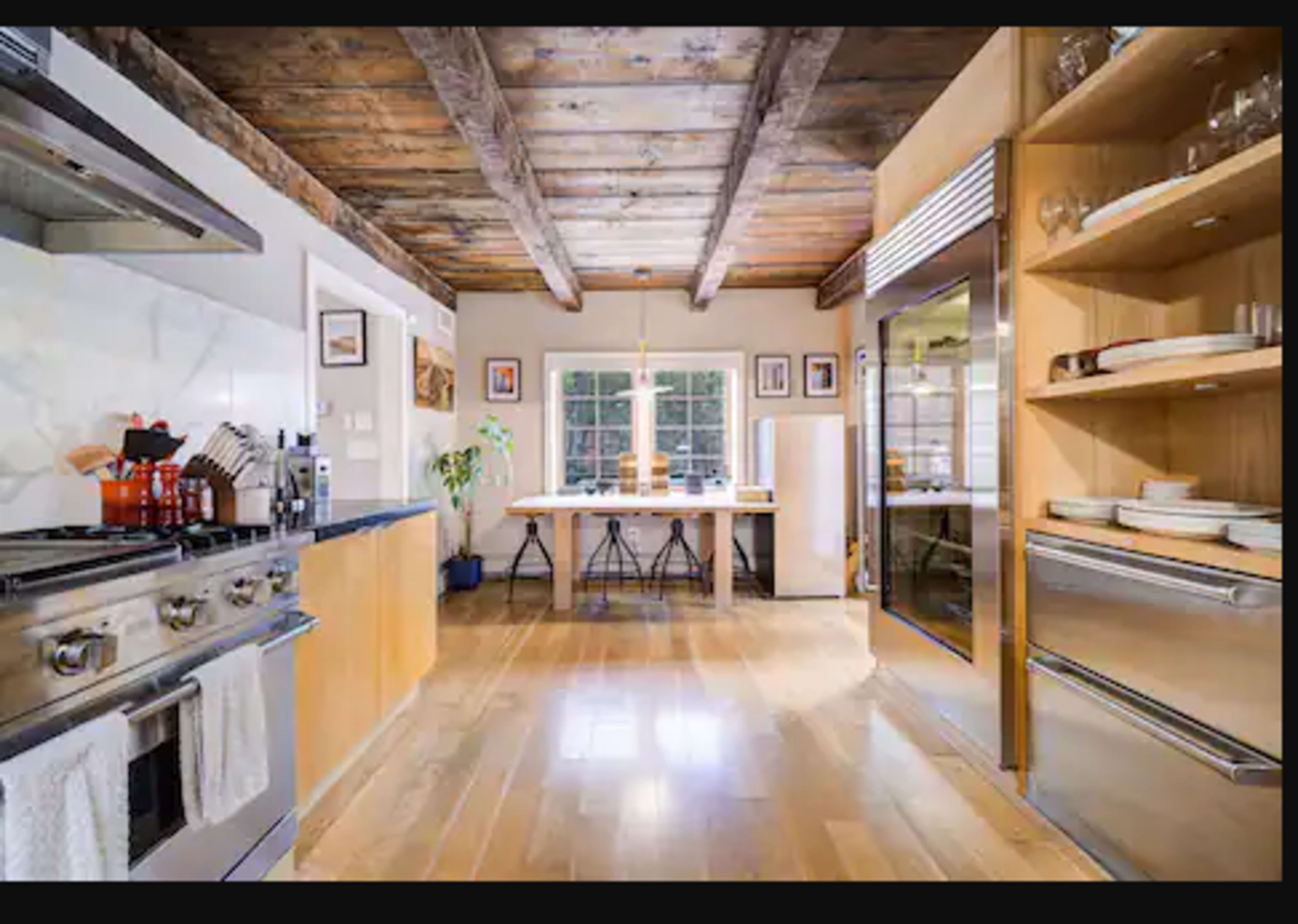A modern kitchen features stainless steel appliances, wooden cabinetry, and a central dining table illuminated by natural light from a large window.