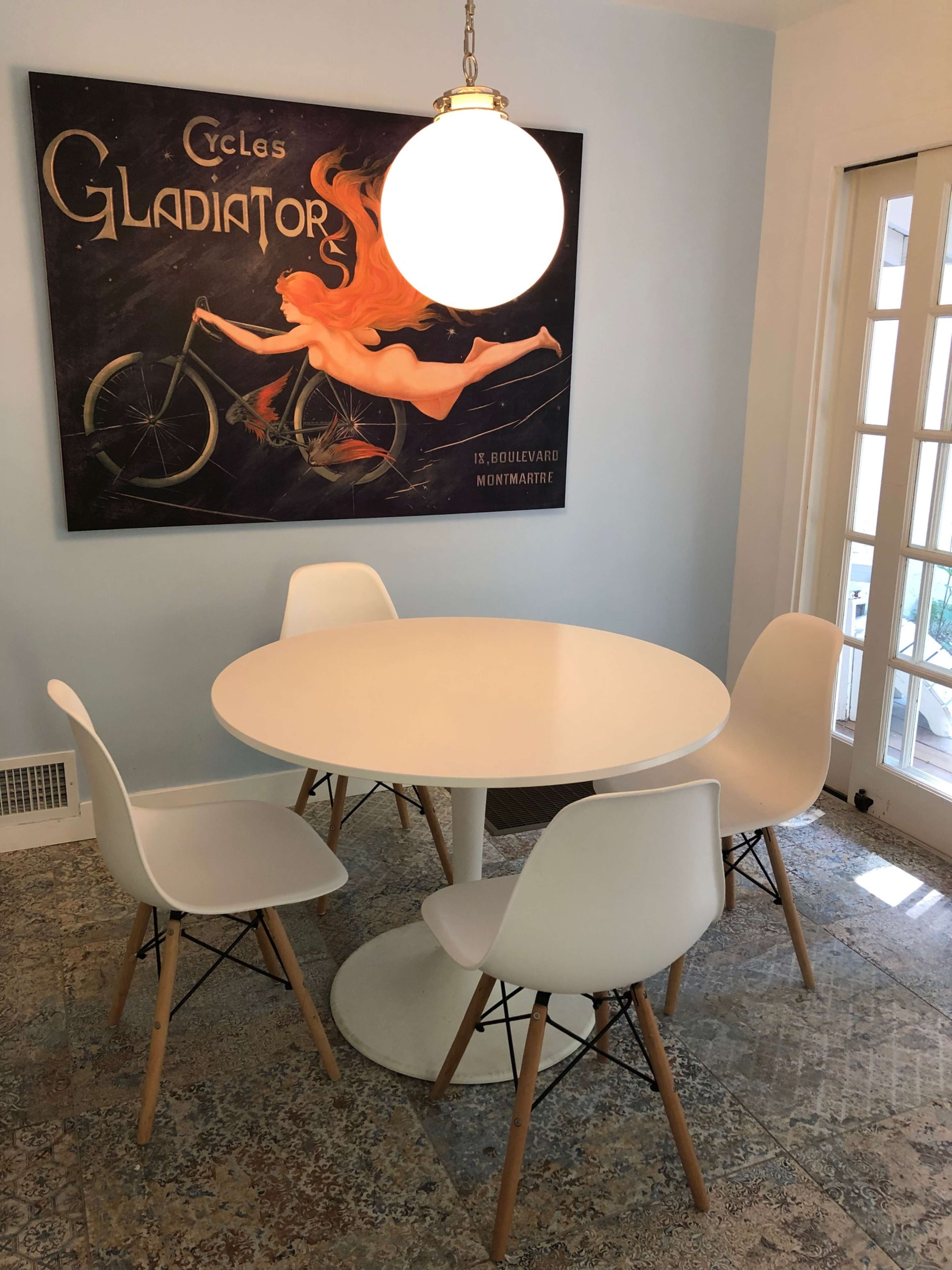 A round white dining table surrounded by four white chairs is positioned beneath a large vintage poster of a woman on a bicycle.