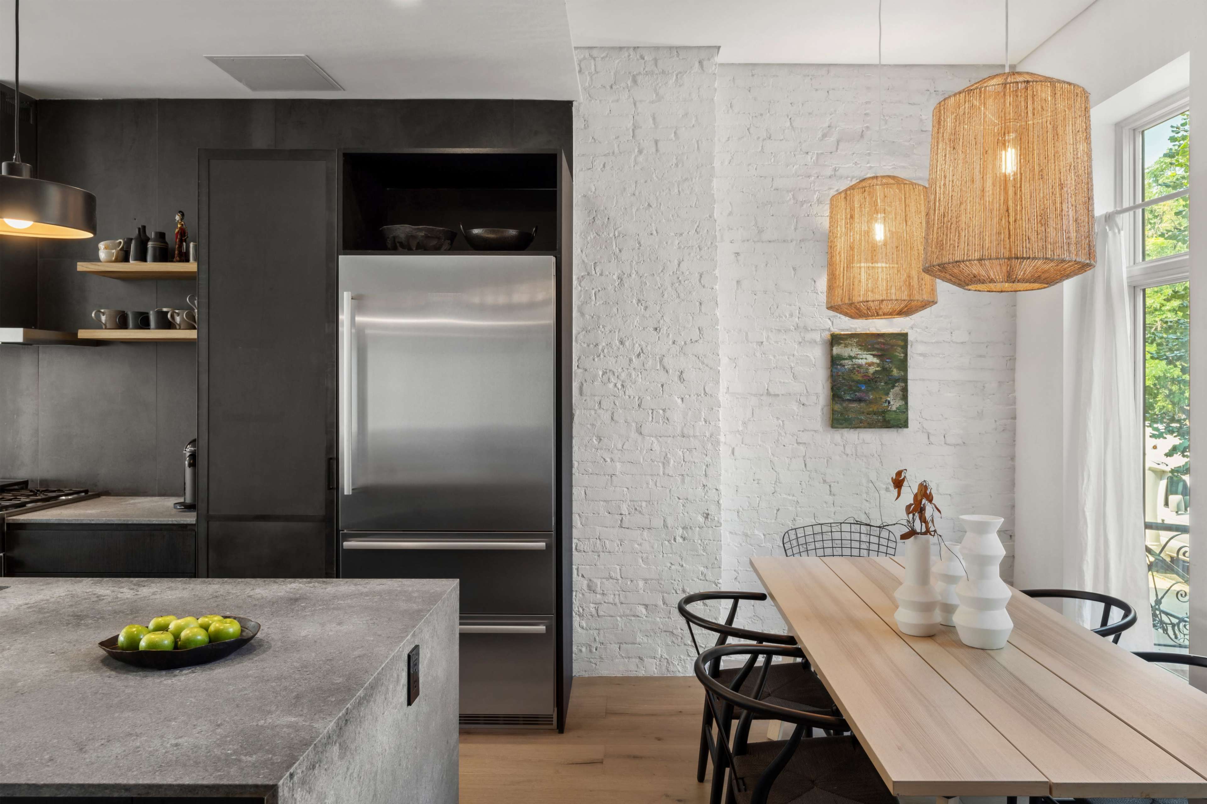 A modern kitchen features a stainless steel refrigerator, a minimalist wooden dining table, and pendant lights above, set against a backdrop of white brick walls.