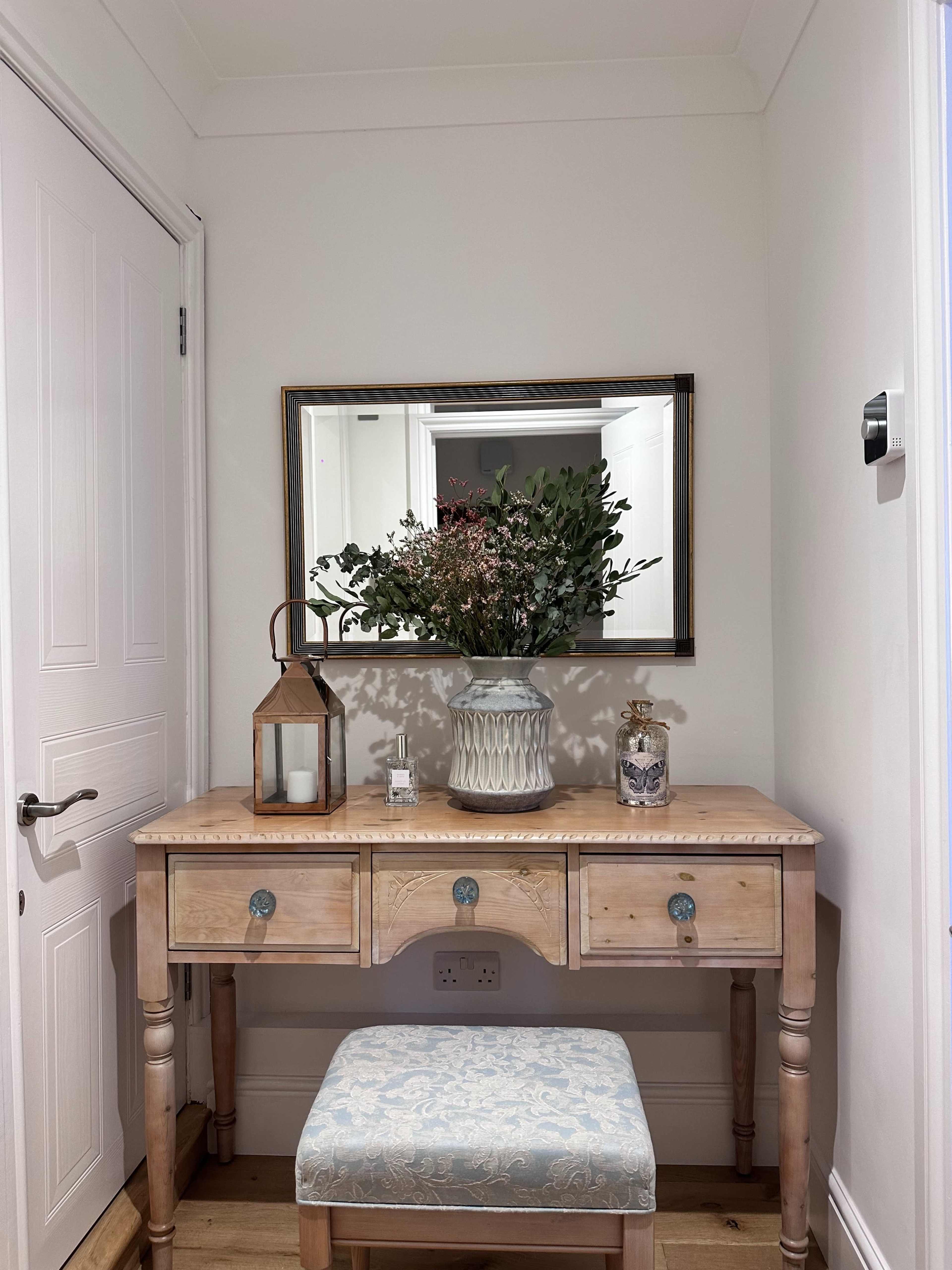 A wooden vanity table with three drawers, a decorative vase, and a lantern sits against a wall, accompanied by a mirror reflecting the arrangement.