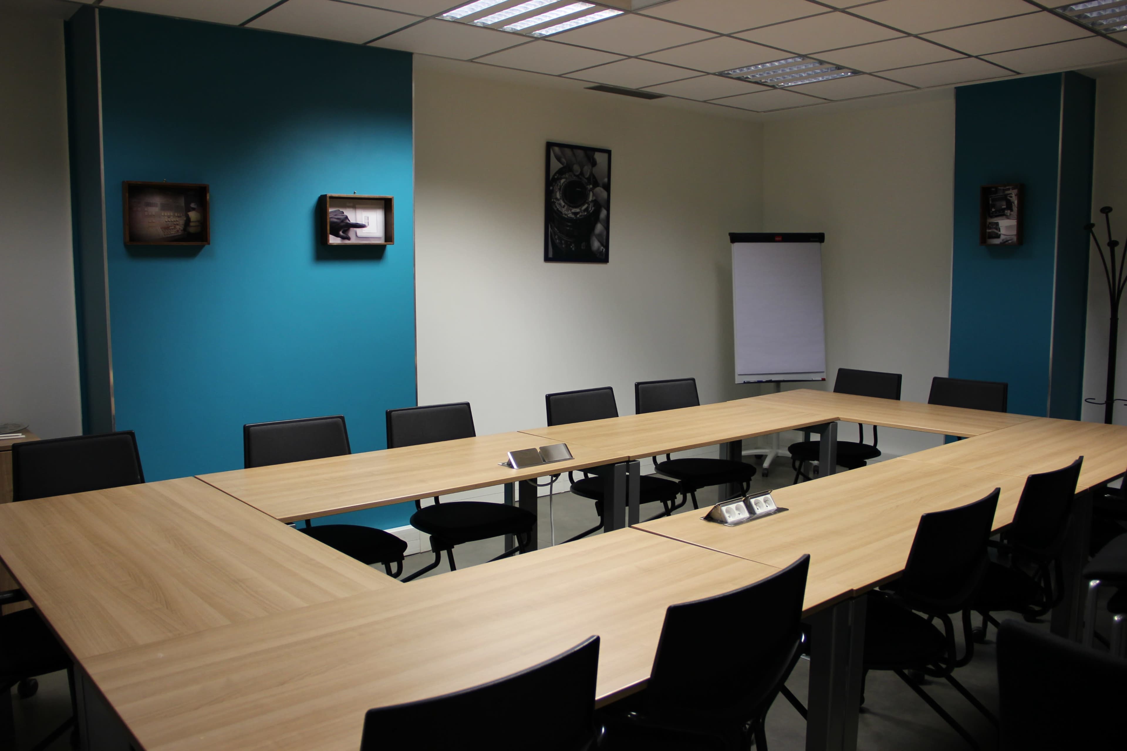 A conference room features a large rectangular table surrounded by black chairs, with teal and white walls, and wall-mounted framed images.