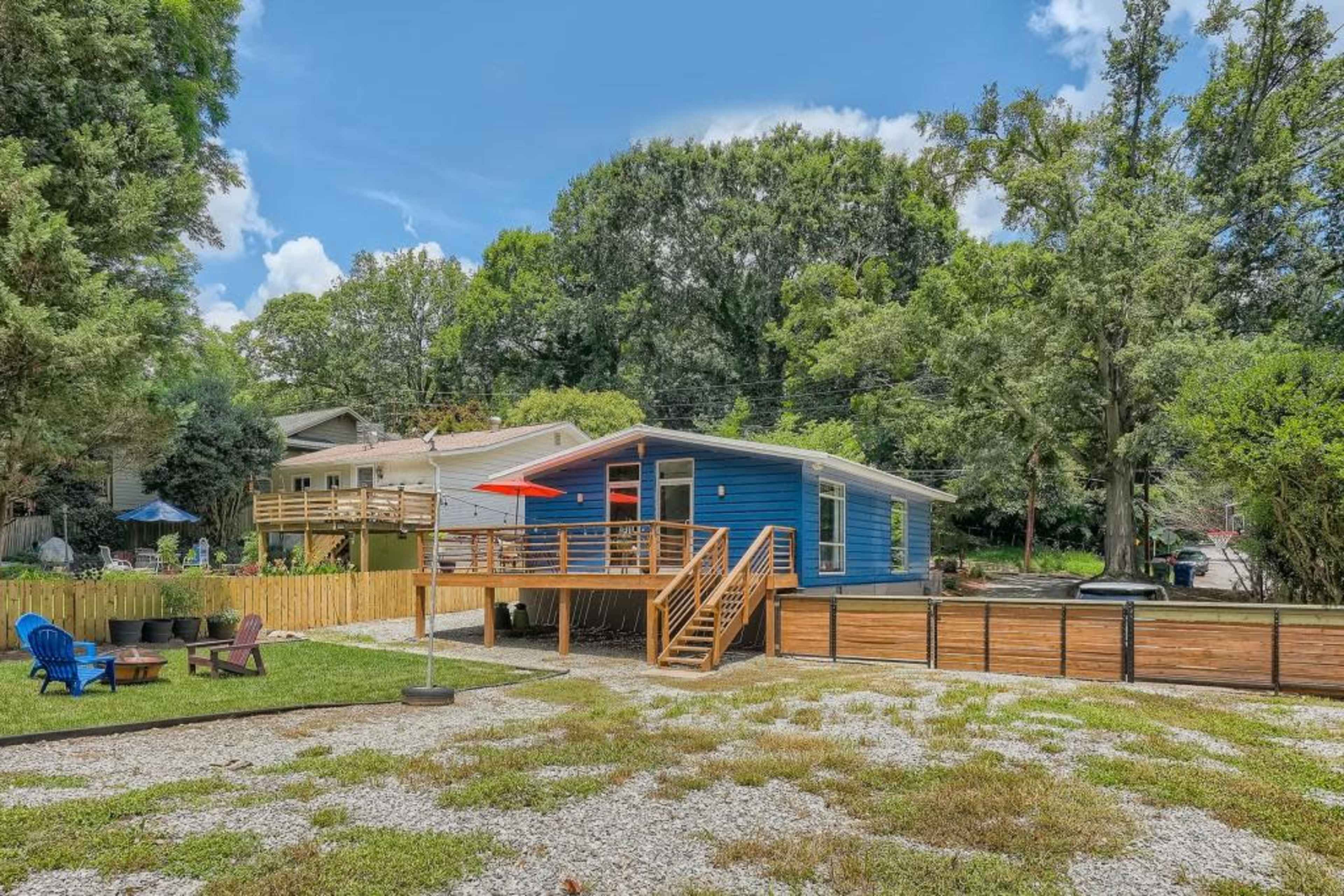 A blue house with a wooden deck and red umbrella is set in a grassy yard surrounded by trees and a gravel area with outdoor seating.