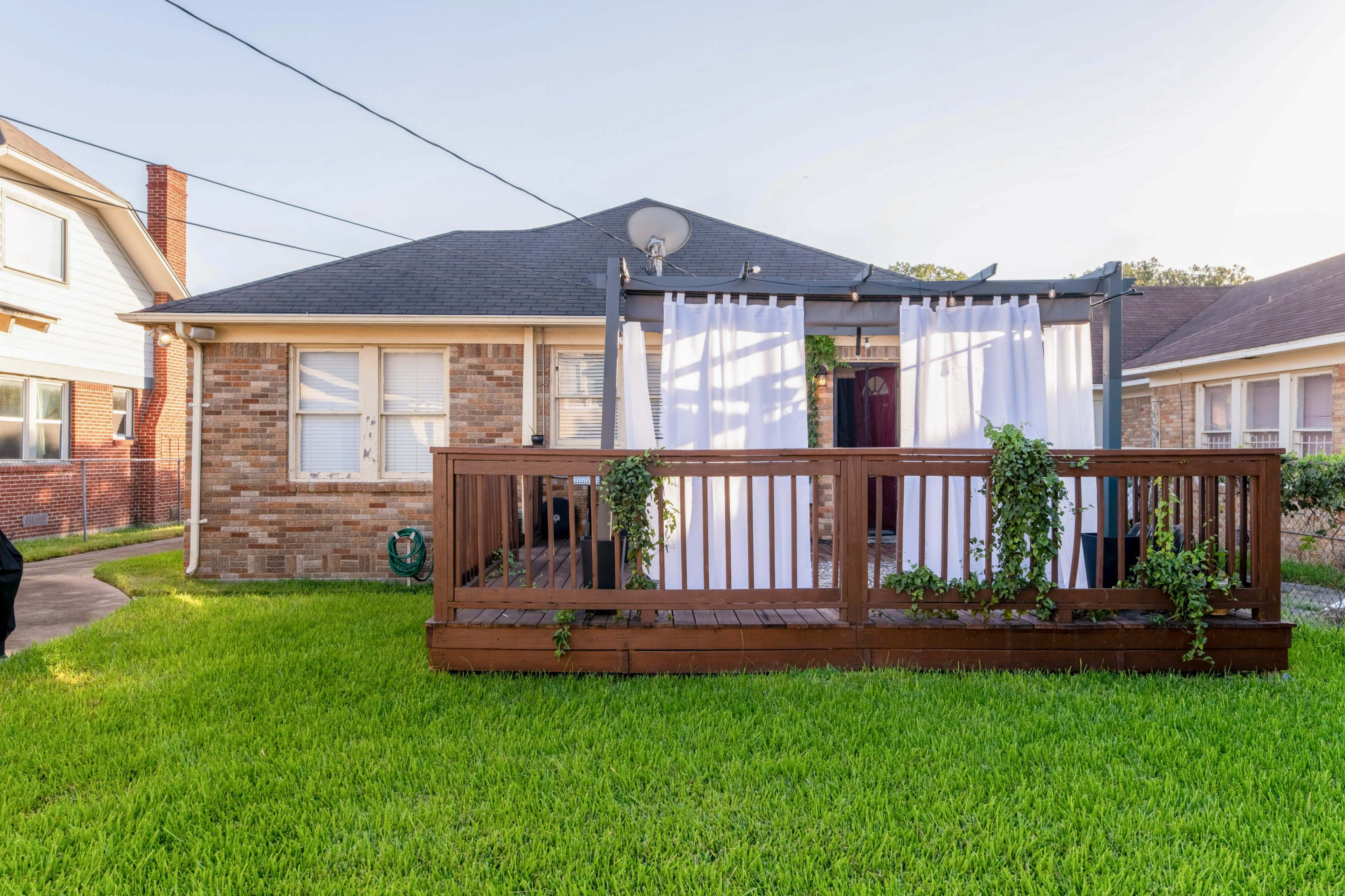 A brick house with a wooden deck is surrounded by green grass and features white curtains hanging from a pergola.