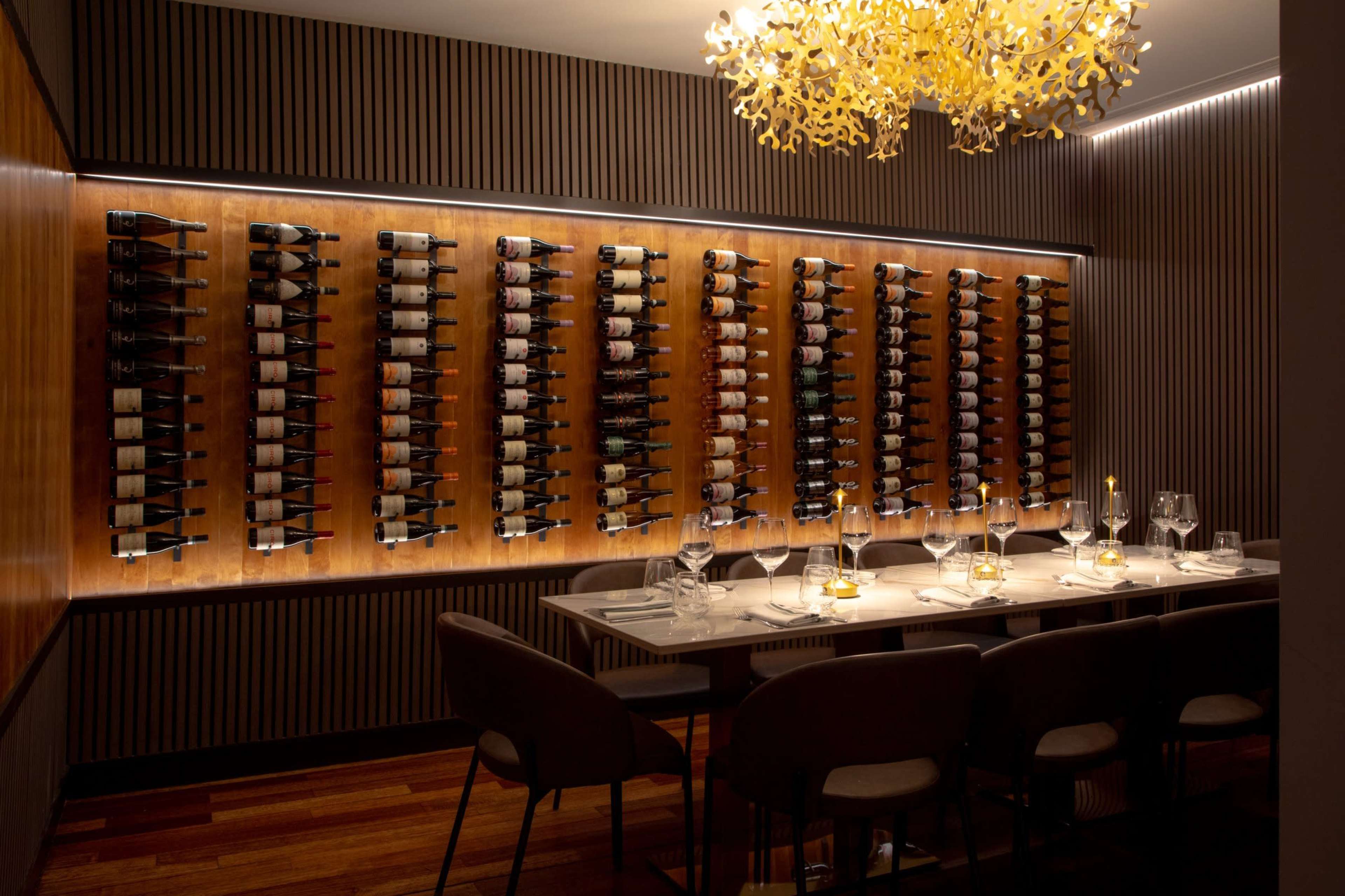 The image shows a modern dining room with a wooden wine rack displaying numerous bottles along one wall, illuminated by soft lighting and featuring a stylish chandelier above a table set for dinner.
