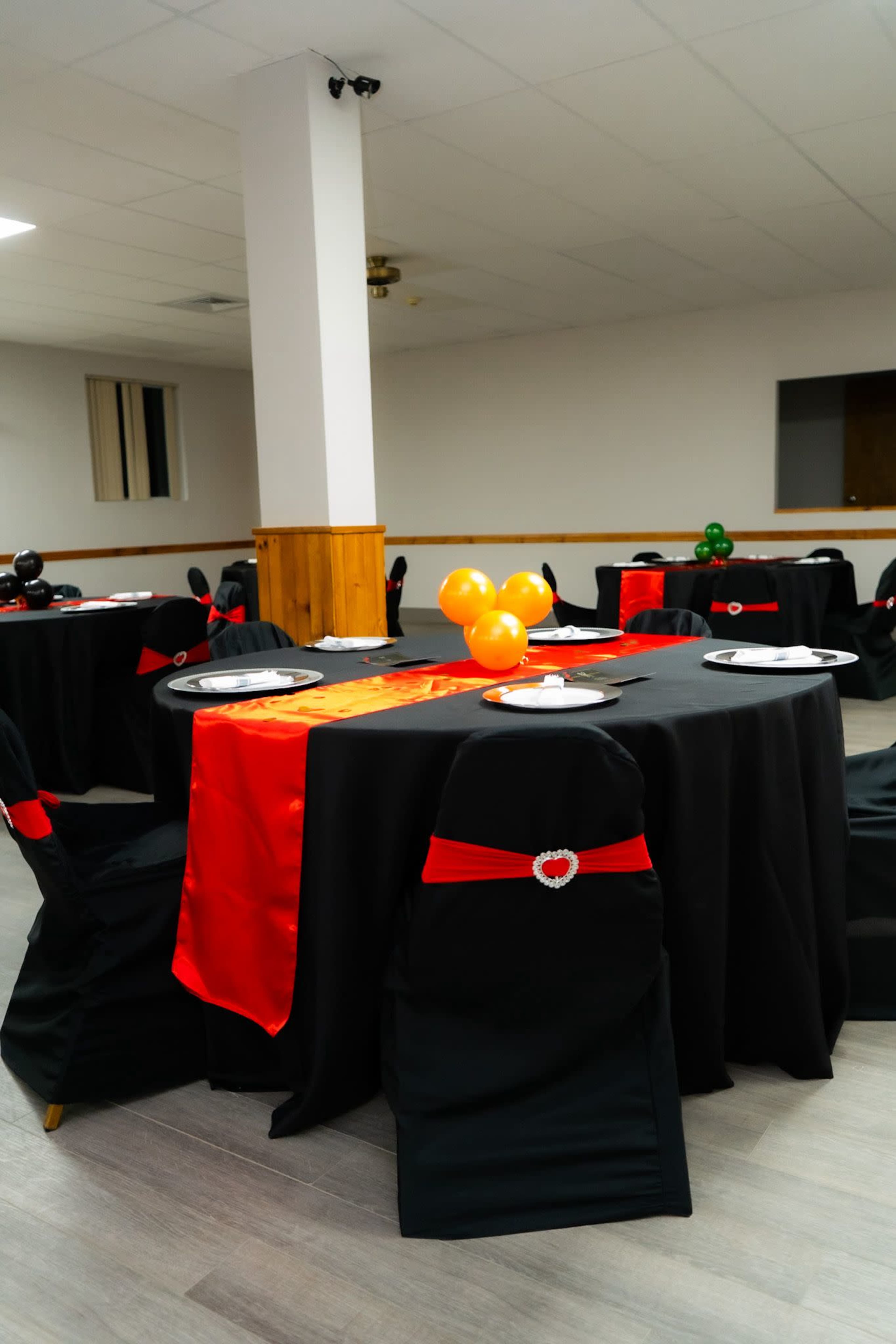 The image shows a banquet room with several tables covered in black tablecloths, accented by red sashes on the chairs and balloons arranged on the tables.
