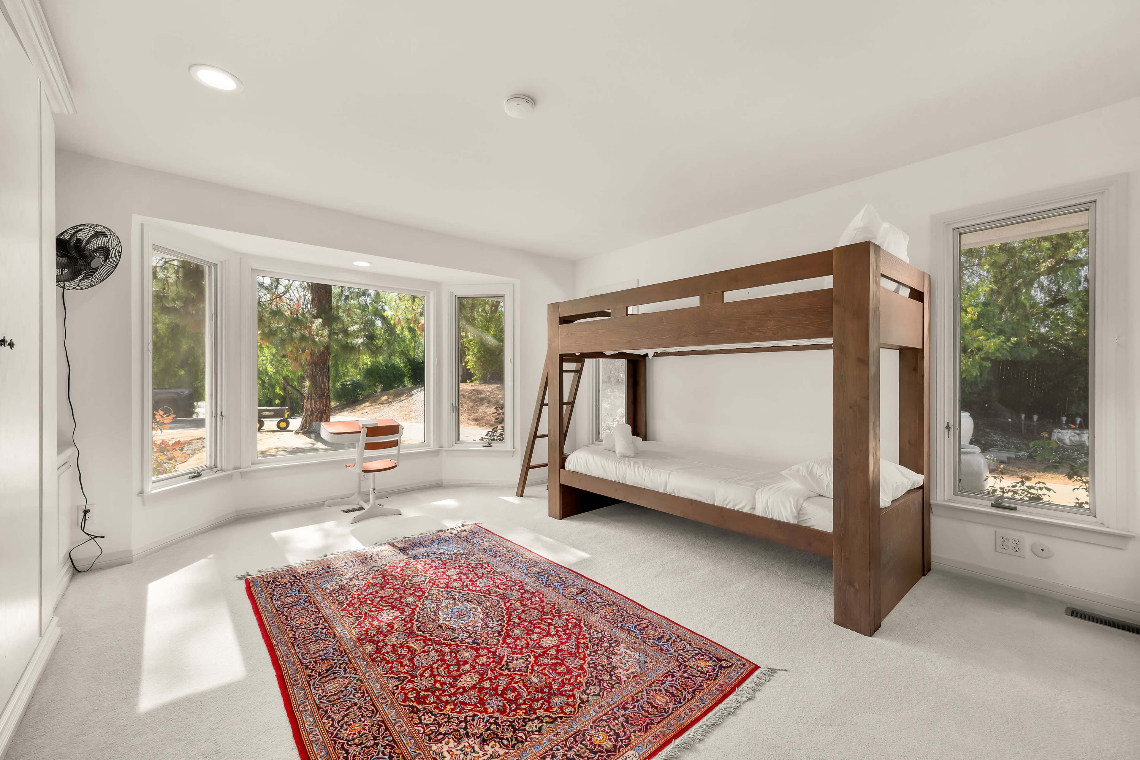 The image depicts a bright room with a wooden bunk bed, a red patterned rug, and large windows allowing natural light.