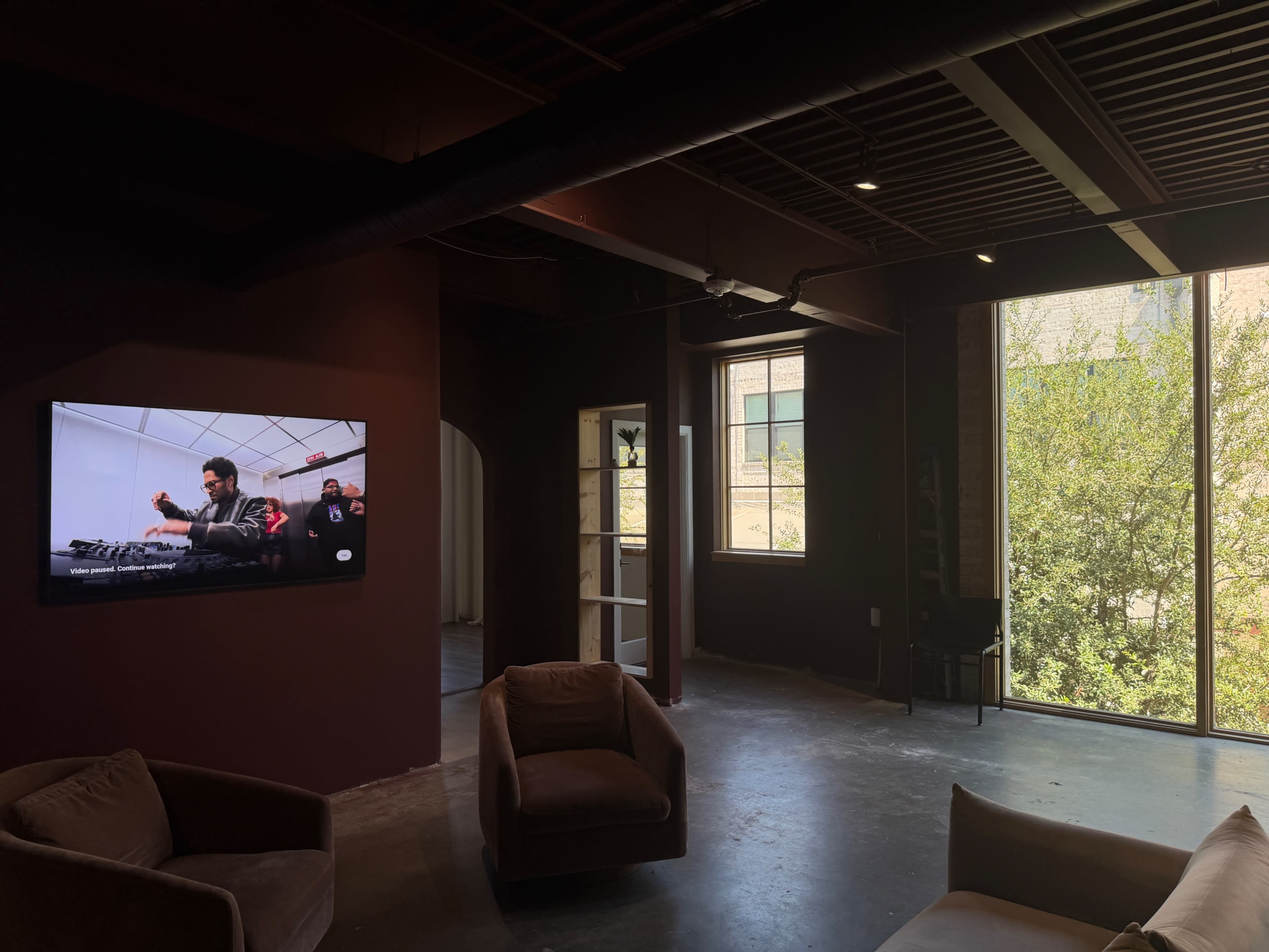 The scene depicts a room with a television displaying a DJ performance, two large windows allowing natural light in, and seating arranged in the foreground.