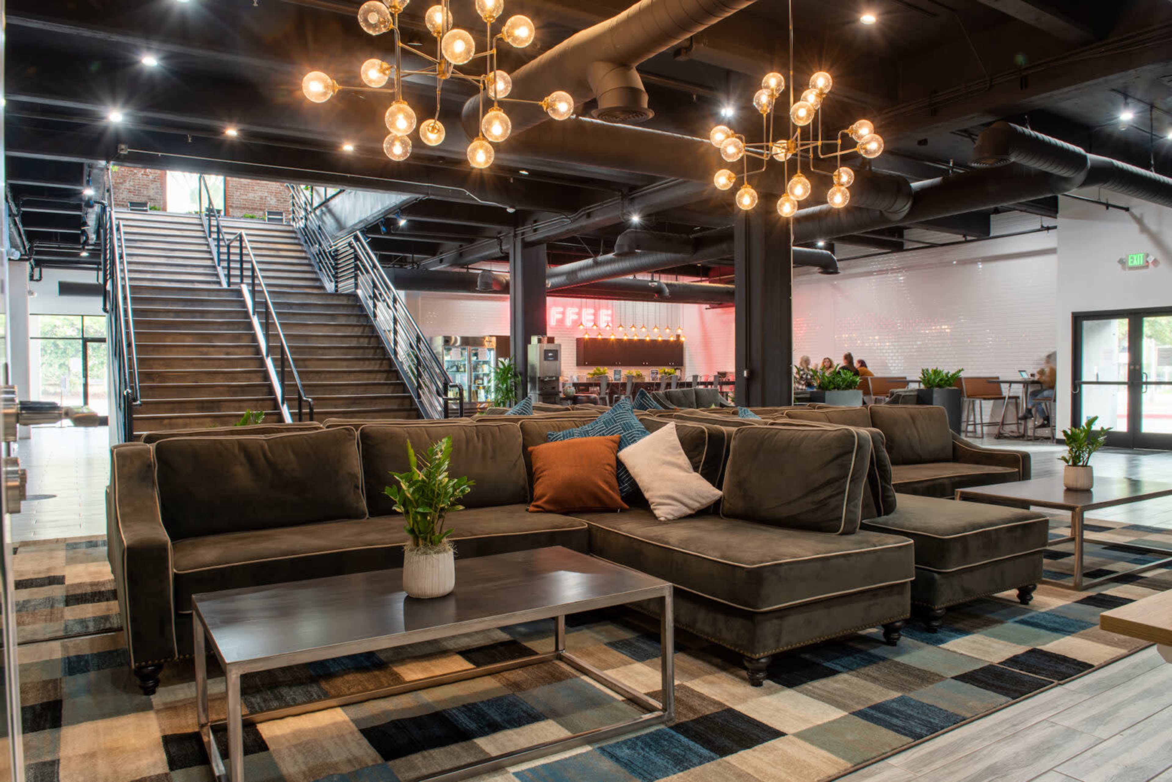The image shows a modern lobby area with a large sectional sofa, decorative lighting fixtures, a coffee table, and a staircase in the background.