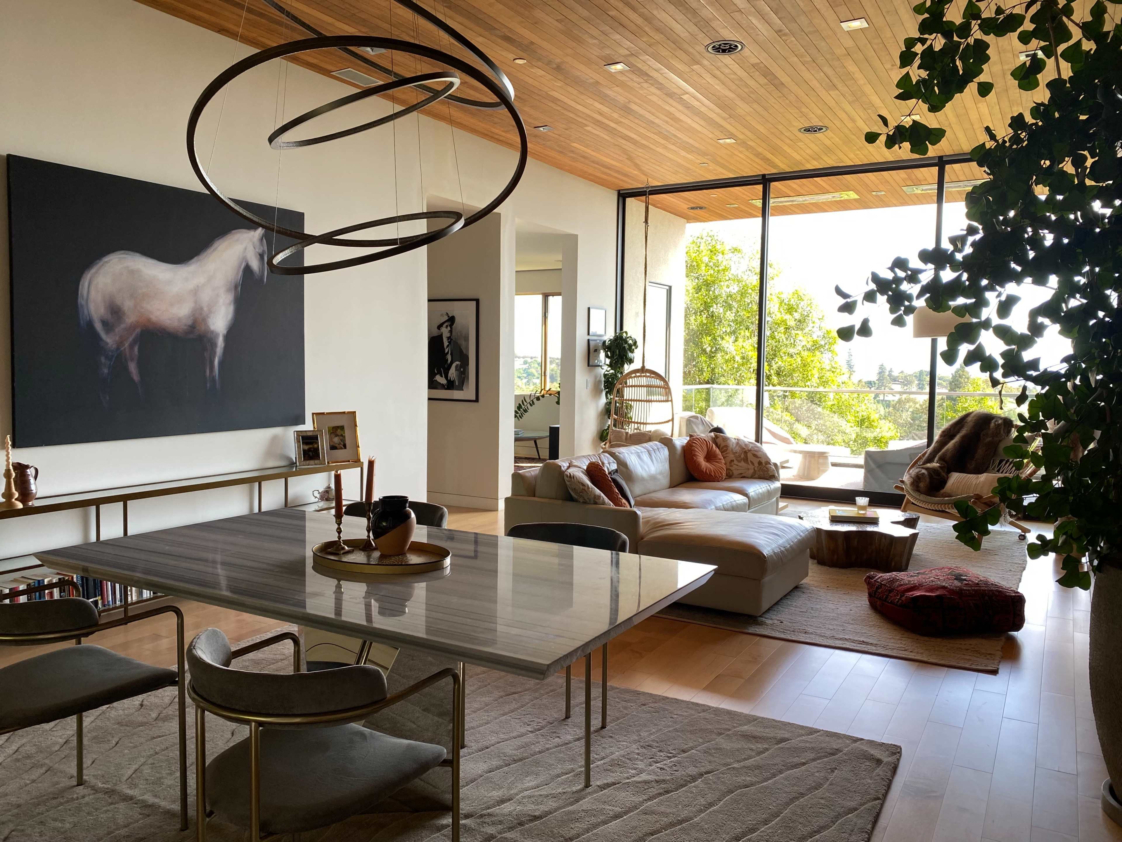 A modern living space features a dining area with a marble table and chairs, a comfortable lounge with a leather sofa, and a large artwork of a horse on the wall, all surrounded by large windows and greenery.