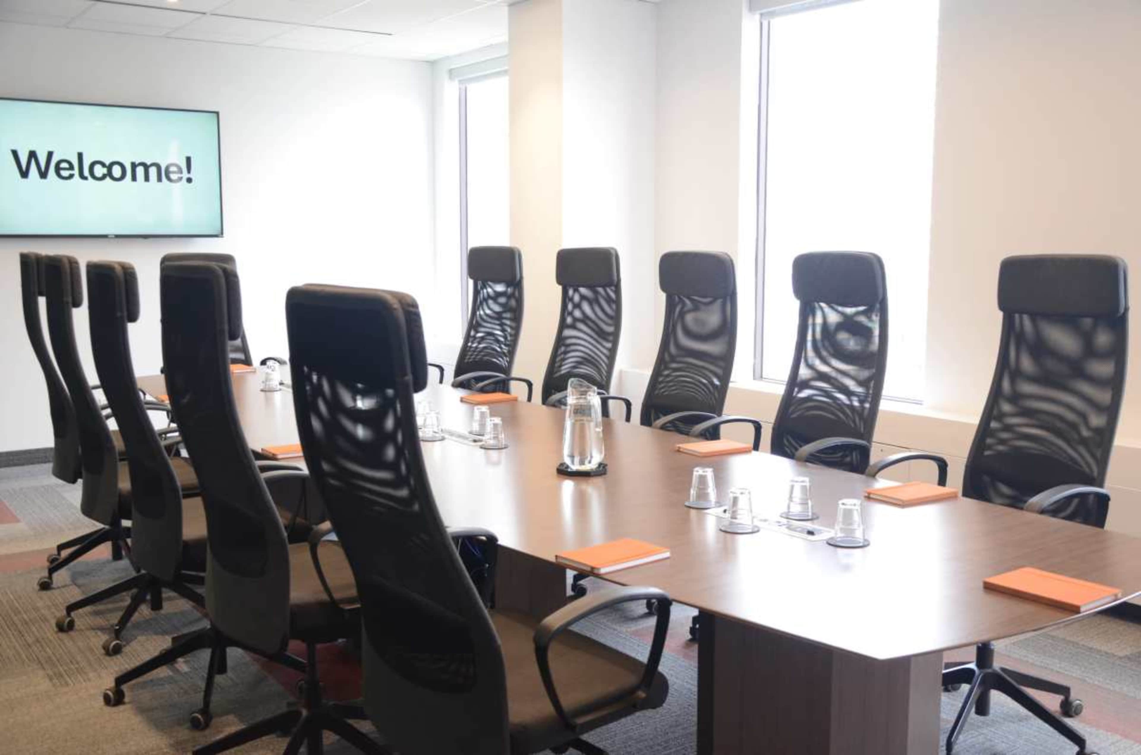 A modern conference room features a large wooden table surrounded by eight high-backed ergonomic chairs, with a welcome display on the wall.